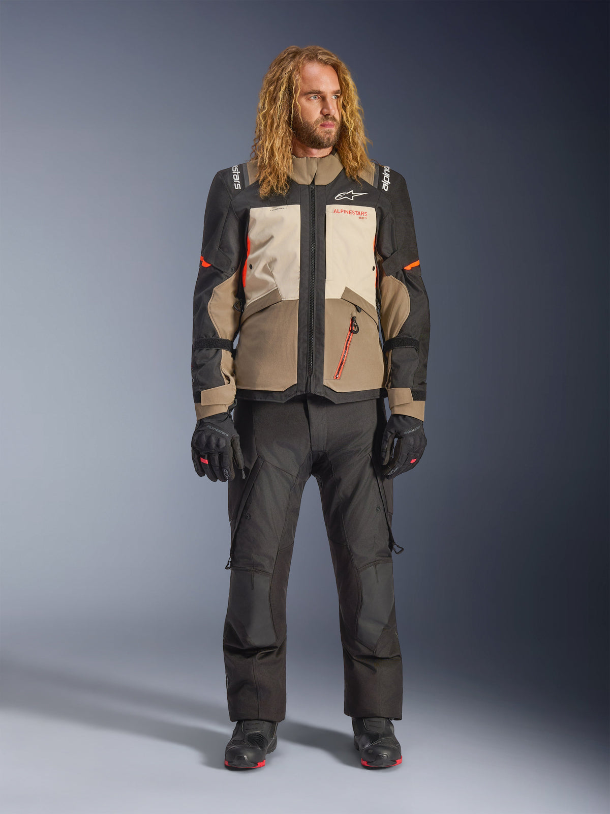 Alpinestars Andes V4 Drystar® Jacke, Walnut Mastic Black, getragen mit schwarzer Motorradhose und Handschuhen, im mehrteiligen Adventure-Design mit orangefarbenen Akzenten und Alpinestars-Branding, technische Textilausrüstung für Fahrten bei jedem Wetter