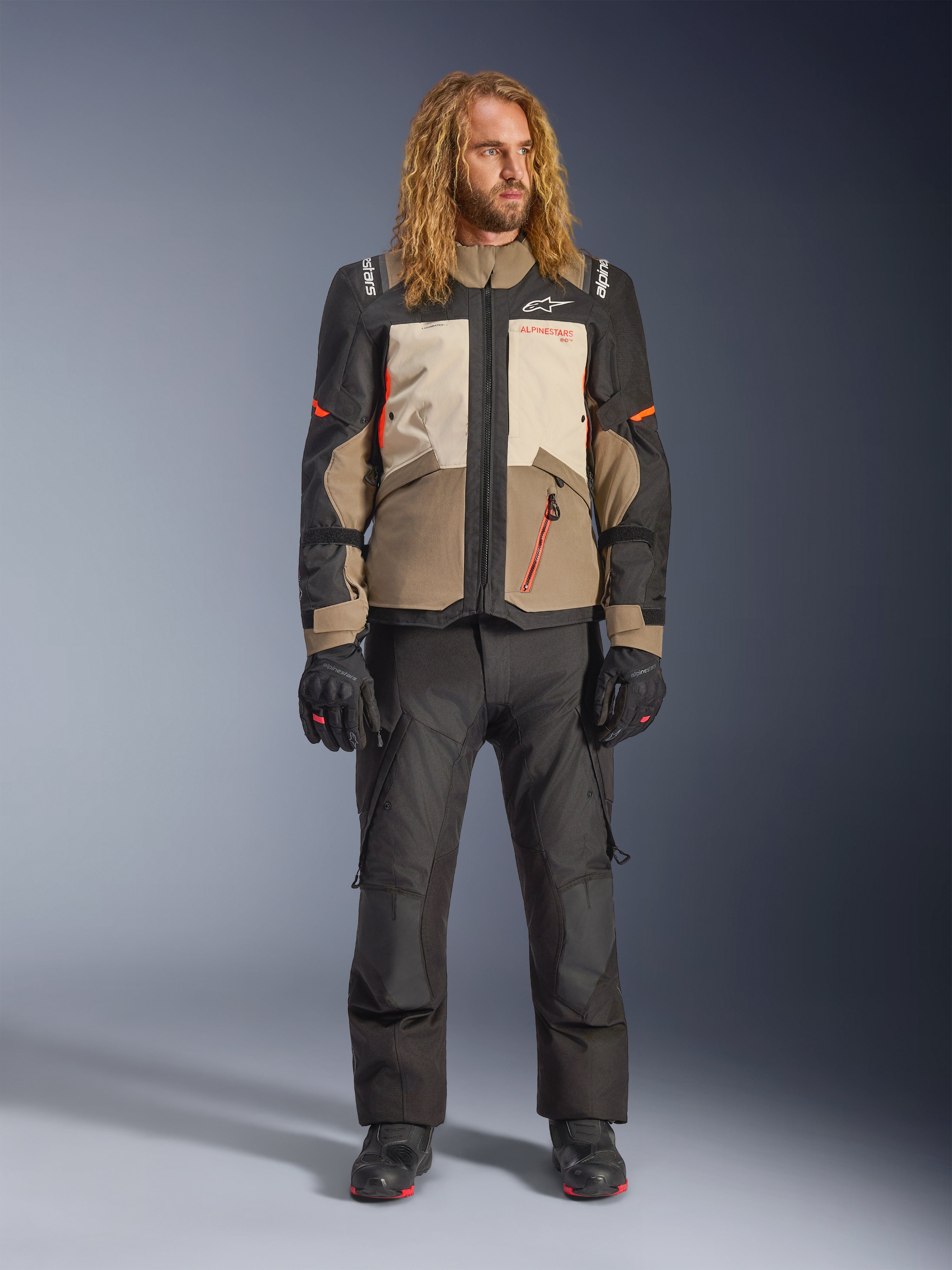 Alpinestars Andes V4 Drystar® Jacke, Walnut Mastic Black, getragen mit schwarzer Motorradhose und Handschuhen, im mehrteiligen Adventure-Design mit orangefarbenen Akzenten und Alpinestars-Branding, technische Textilausrüstung für Fahrten bei jedem Wetter