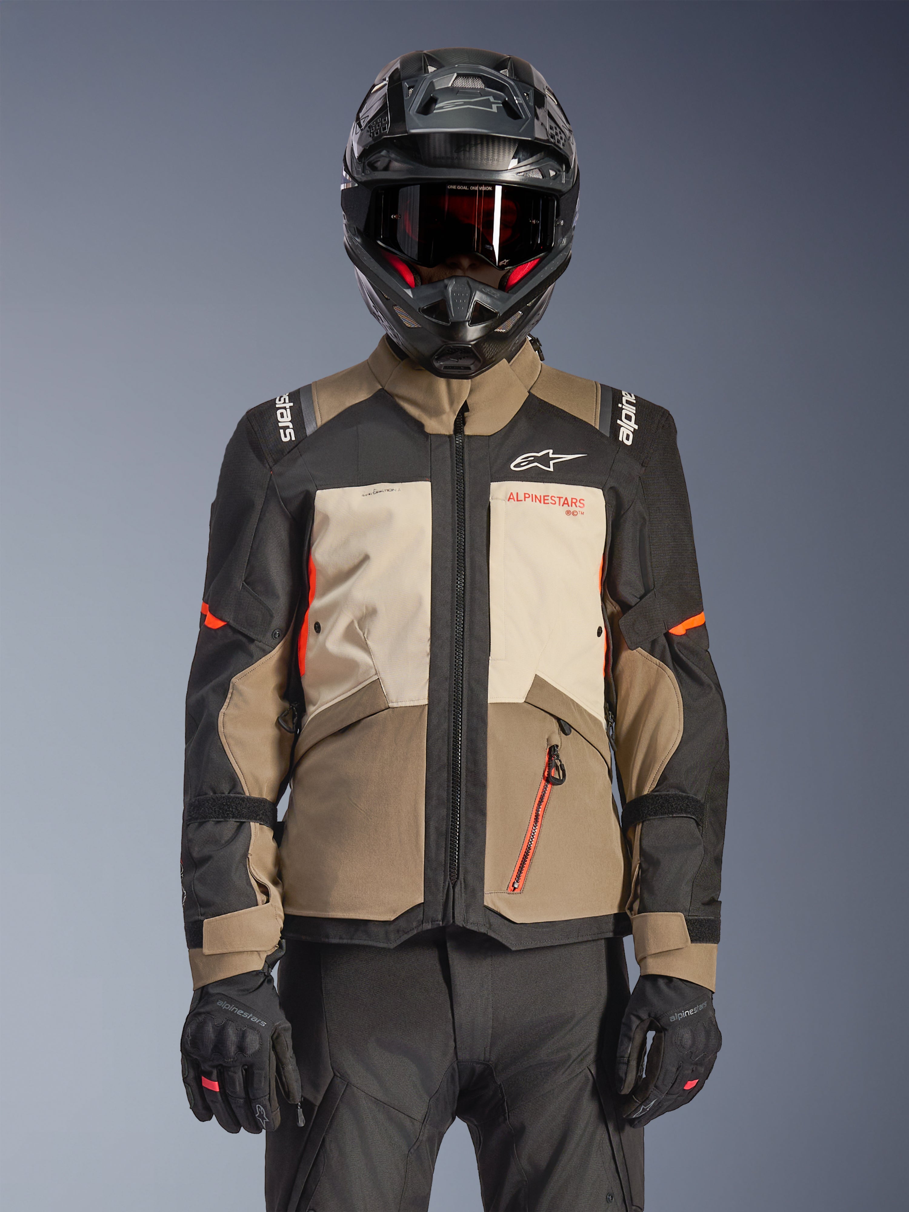 Alpinestars Andes V4 Drystar® Jacke, Walnut Mastic Black, Adventure-Touring-Jacke mit walnussbraunen und mastic-cremefarbenen Paneelen, schwarzen Ärmeln und leuchtend orangefarbenen Akzenten, getragen mit schwarzem Motorradhelm, dunkler Brille und schwarzen Handschuhen