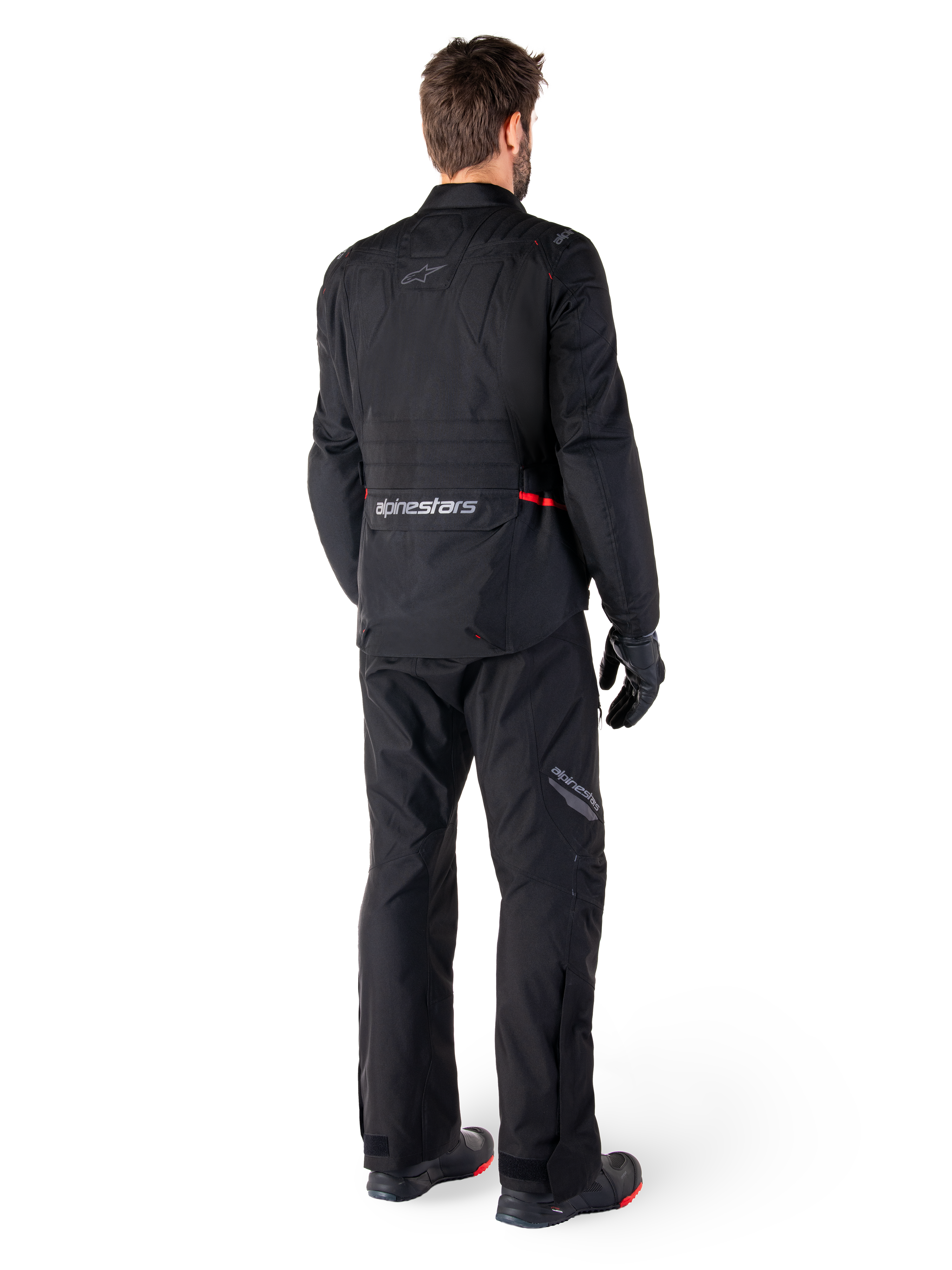 Rear view of a rider wearing the Alpinestars ST-1 Waterproof Jacket and matching pants in black, featuring subtle red accents, a large lower back cargo pocket with branding, and a streamlined adventure touring design.