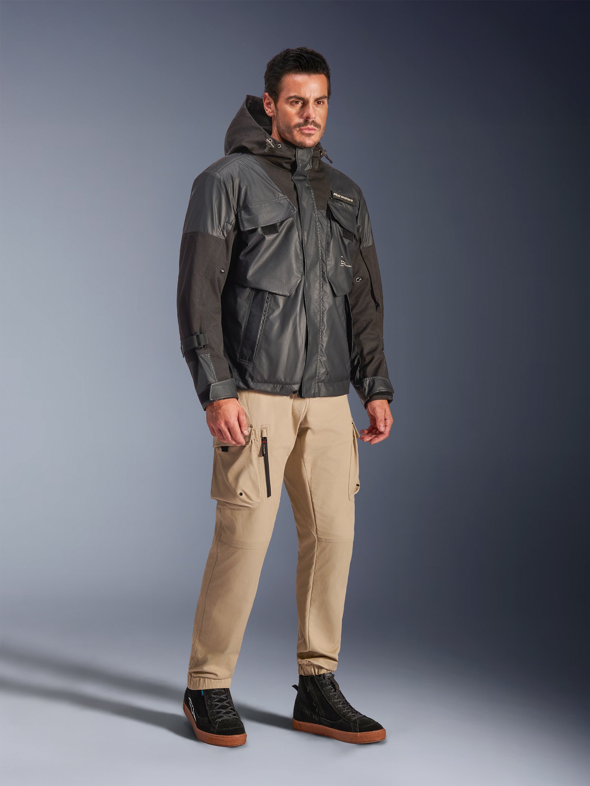 Männliches Modell in der Alpinestars Mospher Waterproof Jacke in Schwarz, wasserdichter Parka im Utility-Look mit Multi-Taschen-Design und Kapuze, kombiniert mit hellbraunen Cargo-Hosen und schwarzen High-Top-Sneakern, urbane Motorrad-Pendlerbekleidung.