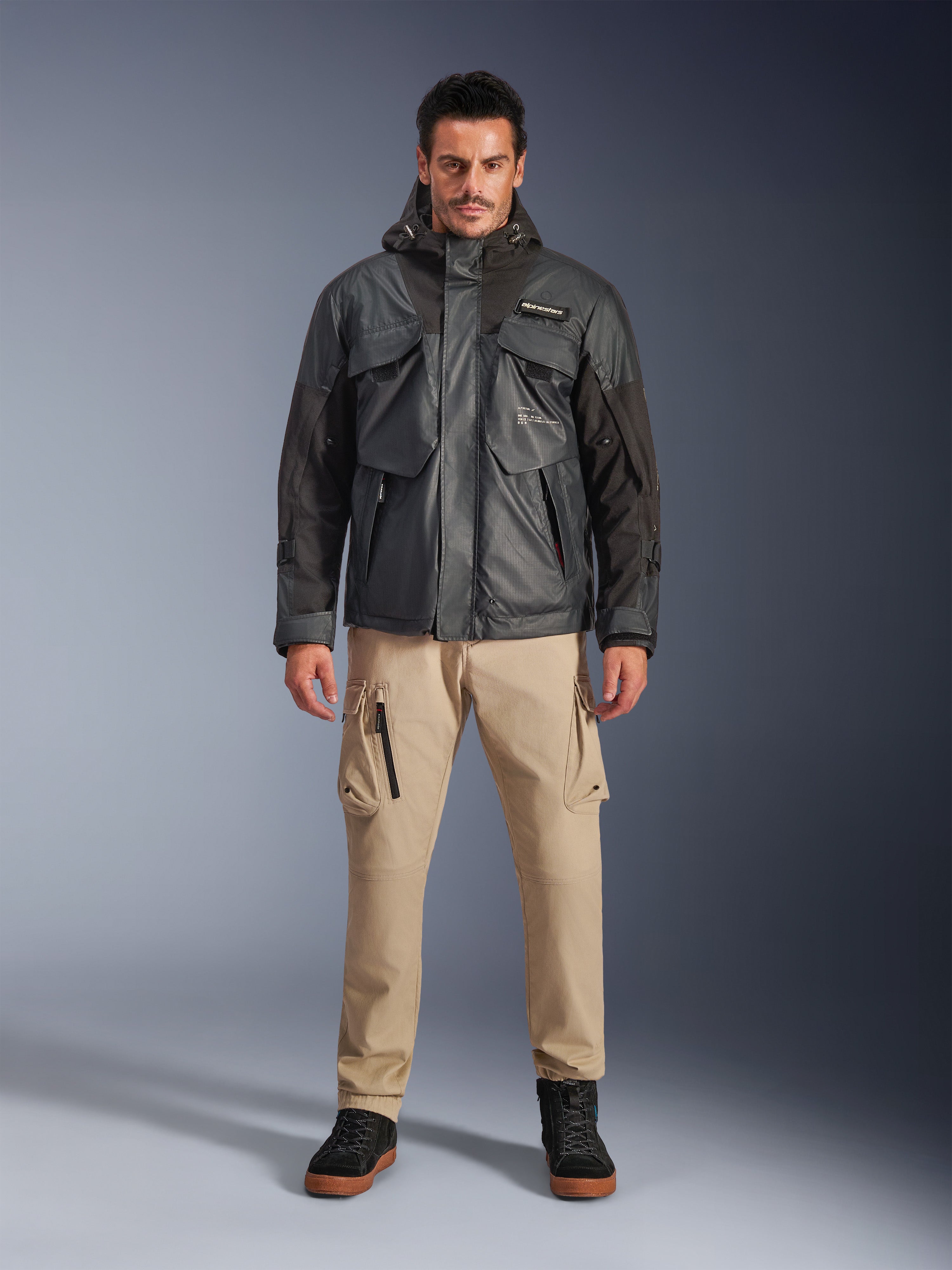 Männliches Model in der Alpinestars Mospher Waterproof Jacke in Schwarz, urbane zeitgemäße Motorradausrüstung, taktisches Utility-Design mit mehreren funktionellen Taschen und Kapuze, kombiniert mit beiger Cargohose und schwarzen Schuhen, Ganzkörper-Studioporträt.