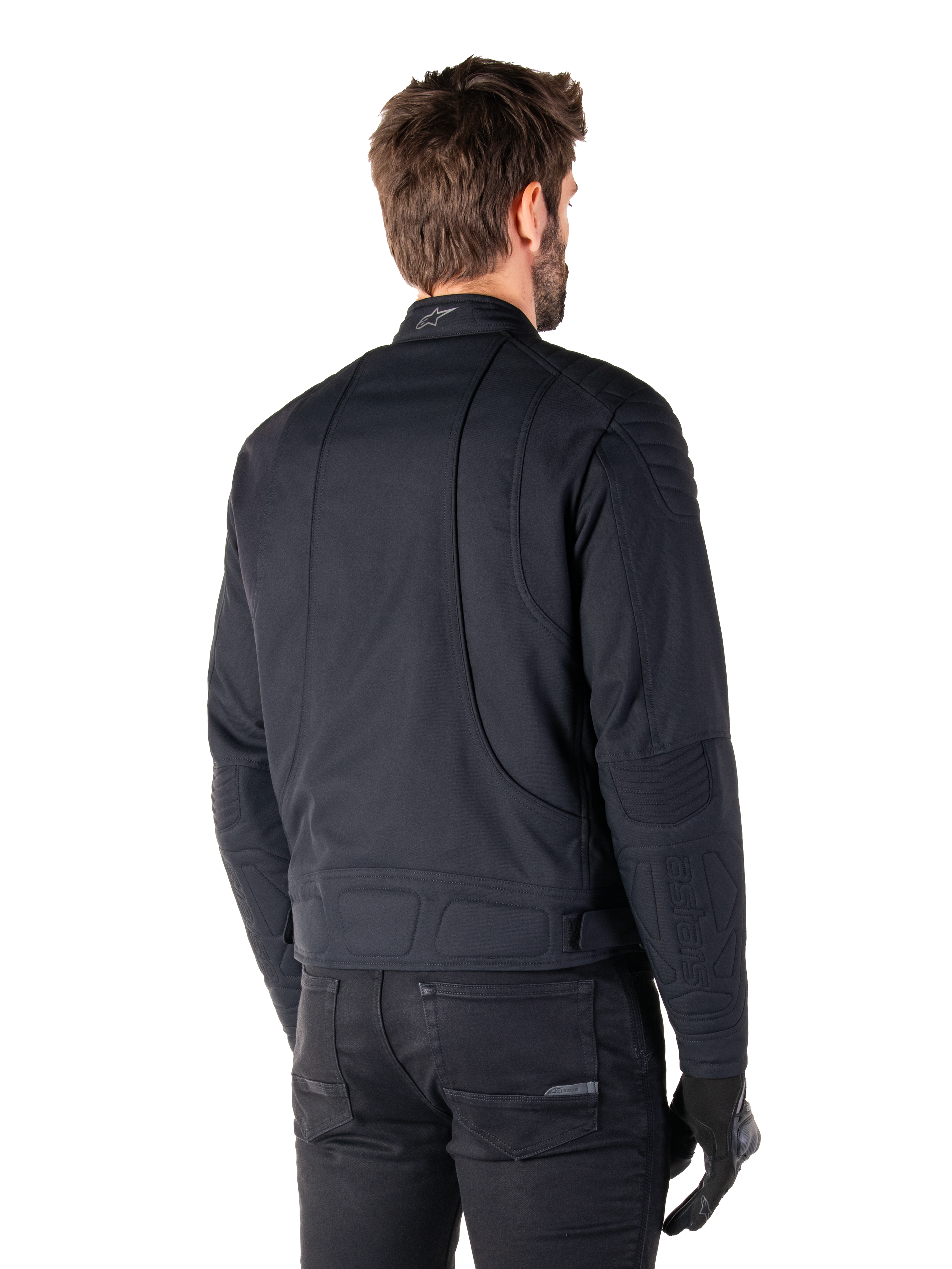 Alpinestars Clayton WR Jacket in Black, rear three-quarter view on a male model, urban motorcycle jacket with quilted padding on shoulders and elbows, technical stitch lines, and reflective logo on the collar, styled with black pants and gloves.