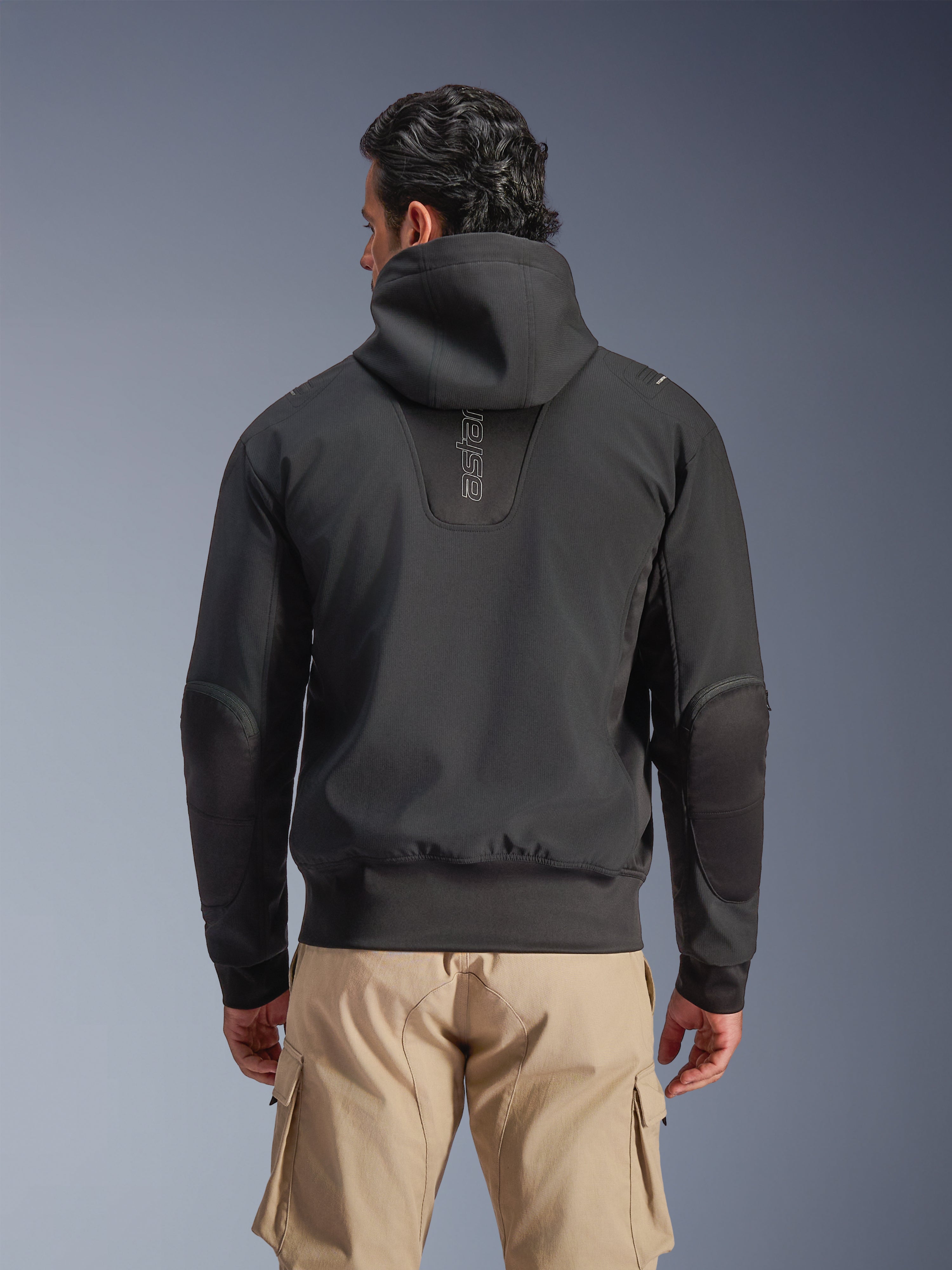 Alpinestars Moblast Wasserdichte Jacke, Schwarz, Rückansicht einer urbanen Motorradjacke mit Kapuze, mit vertikalem 'astars'-Logo auf dem Rücken, elastischem Bund und Bündchen, verstärkten Ellbogen und modernem, technikorientiertem Design, getragen mit einer hellbraunen Cargo-Hose.