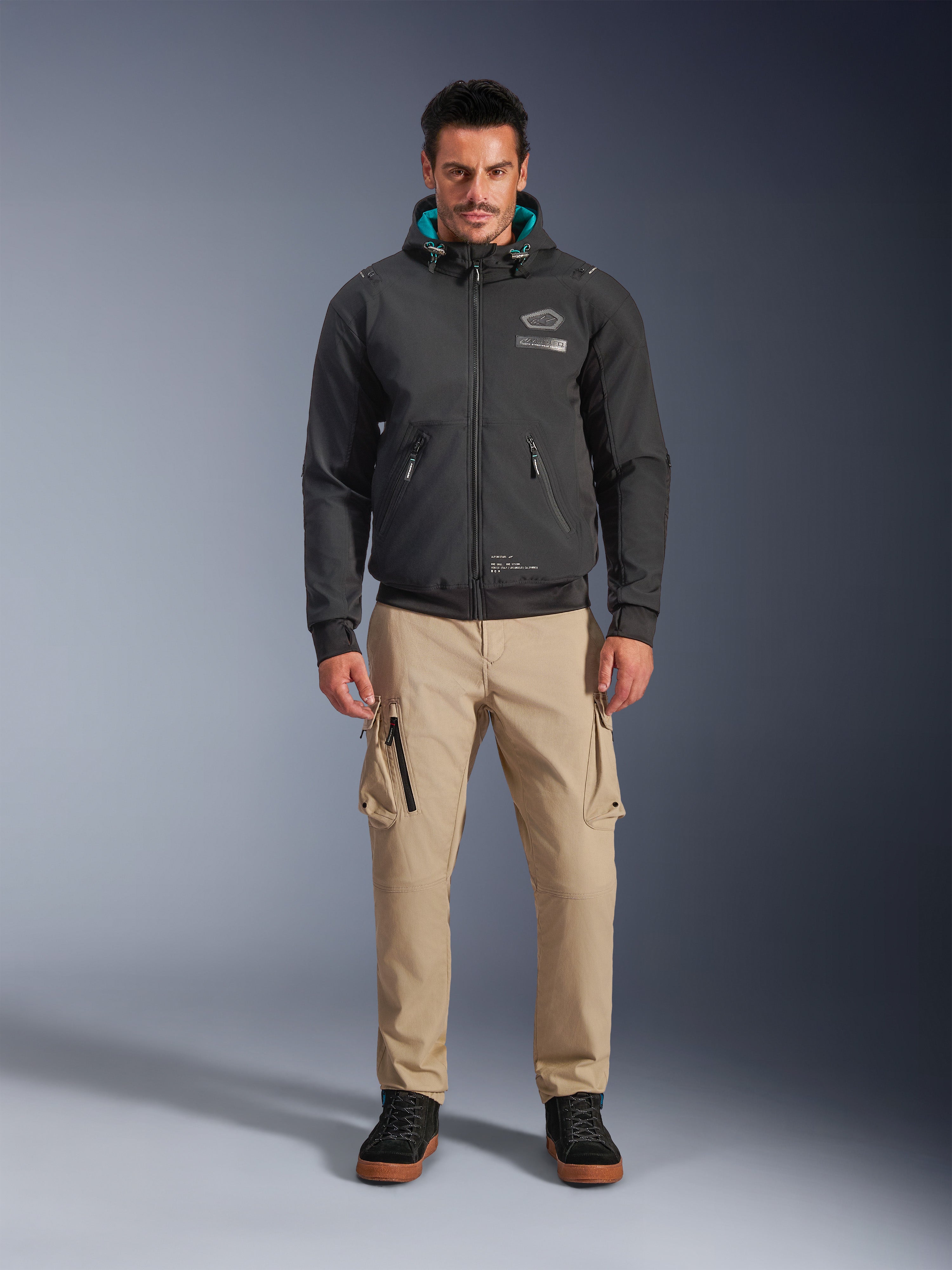 Alpinestars Moblast wasserdichte Jacke, schwarzer urbaner Motorrad-Hoodie mit teal-farbenem Kapuzeninnenfutter, getragen mit sandfarbenen Cargo-Hosen und schwarzen High-Top-Stiefeln, moderne Ganzjahres-Fahrausrüstung
