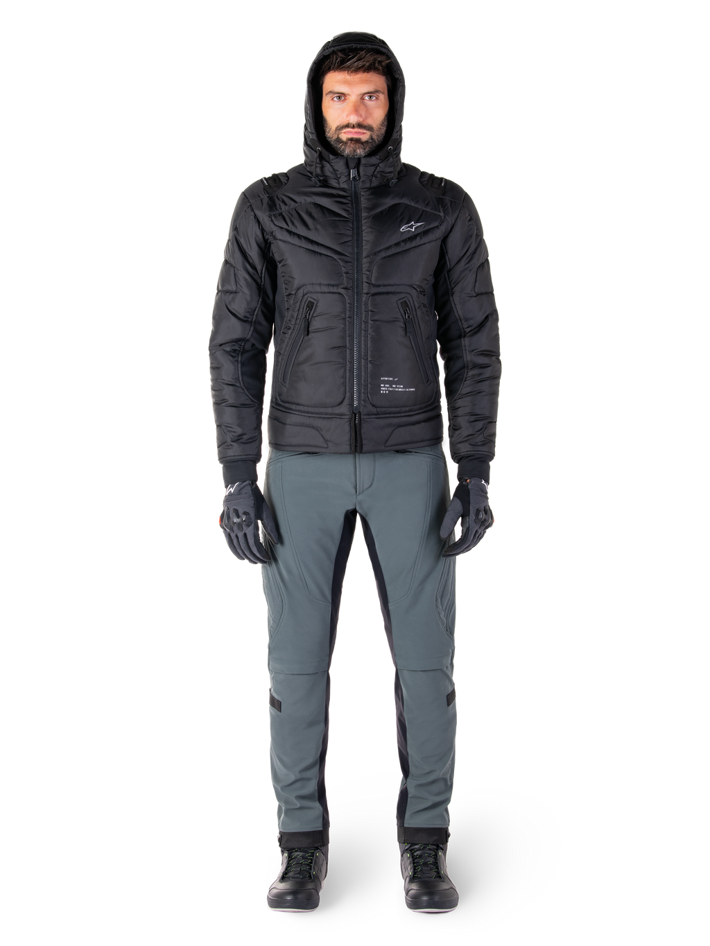 Alpinestars Mohobbs Waterproof Jacket in black, male model wearing quilted padded bomber jacket with hood, paired with grey riding pants, black gloves, and black boots, urban contemporary motorcycle gear.