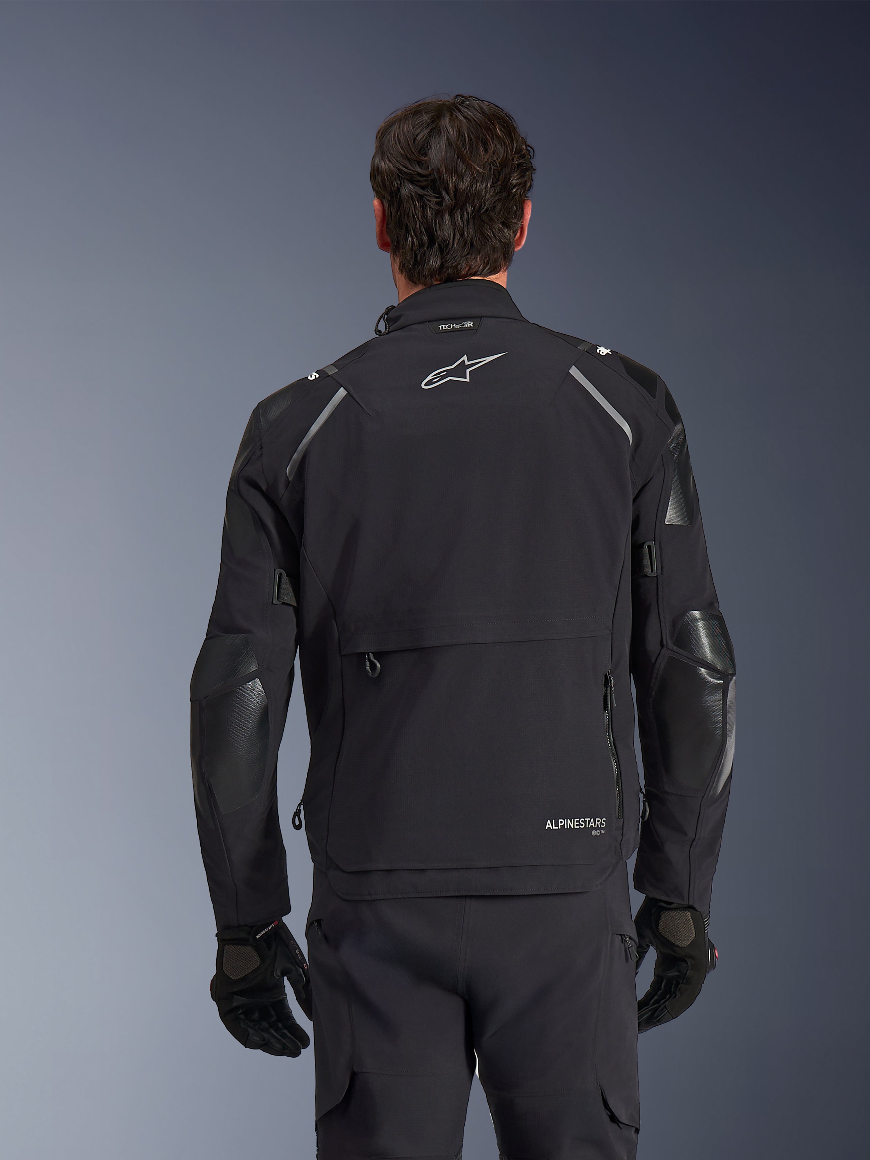 Alpinestars Andes Pro Drystar® XF Jacke, Schwarz und Dunkelgrau, Rückansicht an einem männlichen Modell, mit einem großen hinteren Staufach, reflektierenden Schulterakzenten und Logo-Branding, kombiniert mit schwarzen Handschuhen und einer Hose für Adventure-Touren.