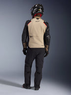 Rider from the back wearing the Alpinestars Andes Pro Drystar® XF Jacket in Walnut Aluminum, tan back panel with black sleeves and red accents, paired with black technical adventure pants and a motorcycle helmet, premium all-weather touring gear.