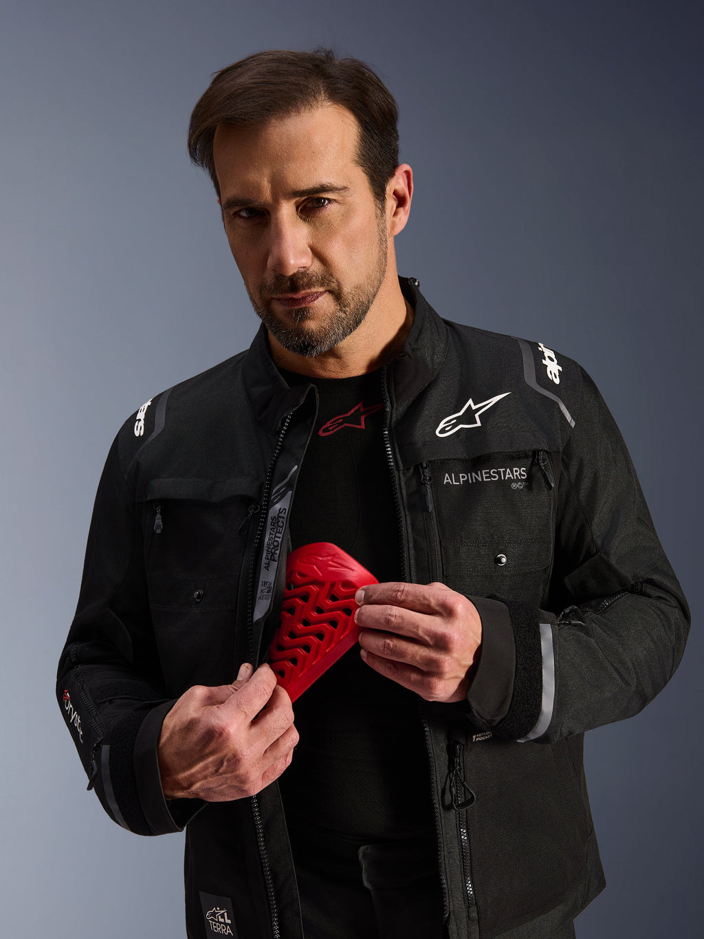 Alpinestars Cusco Drystar® Jacke in Schwarz, männliches Model hält ein rotes Nucleon Flex Pro Protektorenelement, Adventure-Touring-Motorradjacke mit weißem Branding, technische Textilkonstruktion mit Belüftungsreißverschlüssen und mehreren Taschen