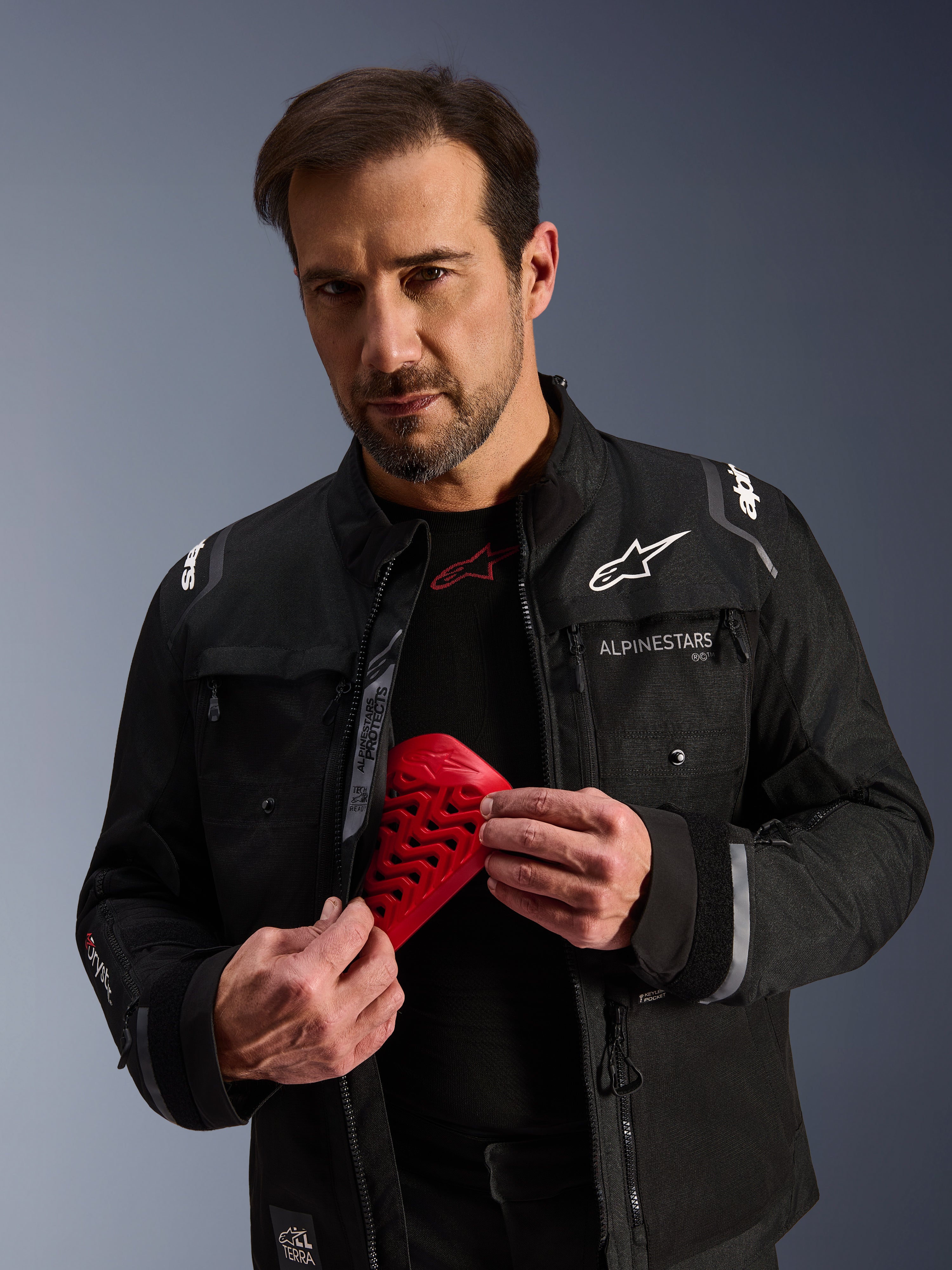 Alpinestars Cusco Drystar® Jacke in Schwarz, männliches Model hält ein rotes Nucleon Flex Pro Protektorenelement, Adventure-Touring-Motorradjacke mit weißem Branding, technische Textilkonstruktion mit Belüftungsreißverschlüssen und mehreren Taschen