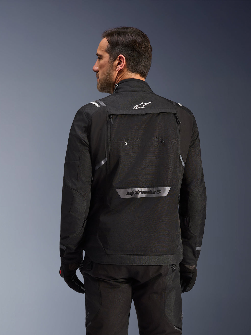 Rückansicht eines Fahrers in der Alpinestars Cusco Drystar® Jacke in Schwarz, eine Textil-Adventure-Tourenjacke mit großer Rückentasche und reflektierendem Branding, kombiniert mit passender schwarzer Hose und Handschuhen