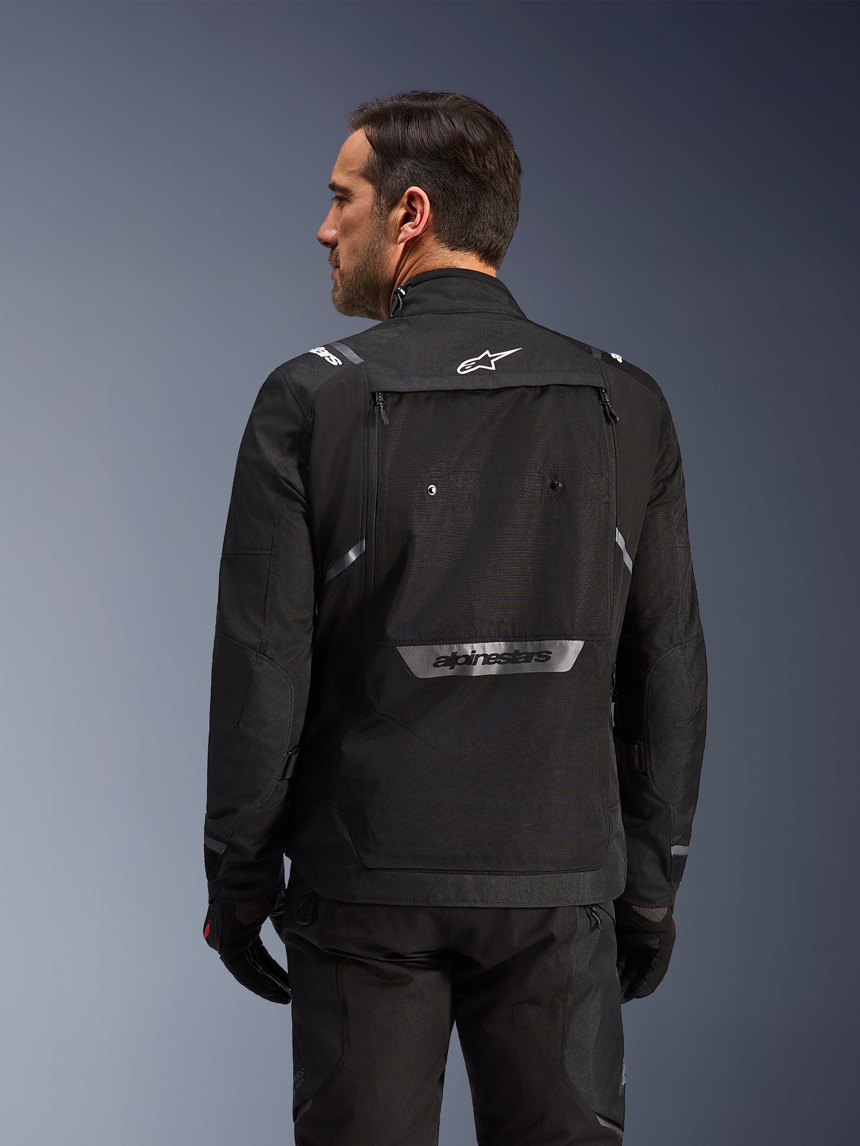 Rückansicht eines Fahrers in der Alpinestars Cusco Drystar® Jacke in Schwarz, eine Textil-Adventure-Tourenjacke mit großer Rückentasche und reflektierendem Branding, kombiniert mit passender schwarzer Hose und Handschuhen