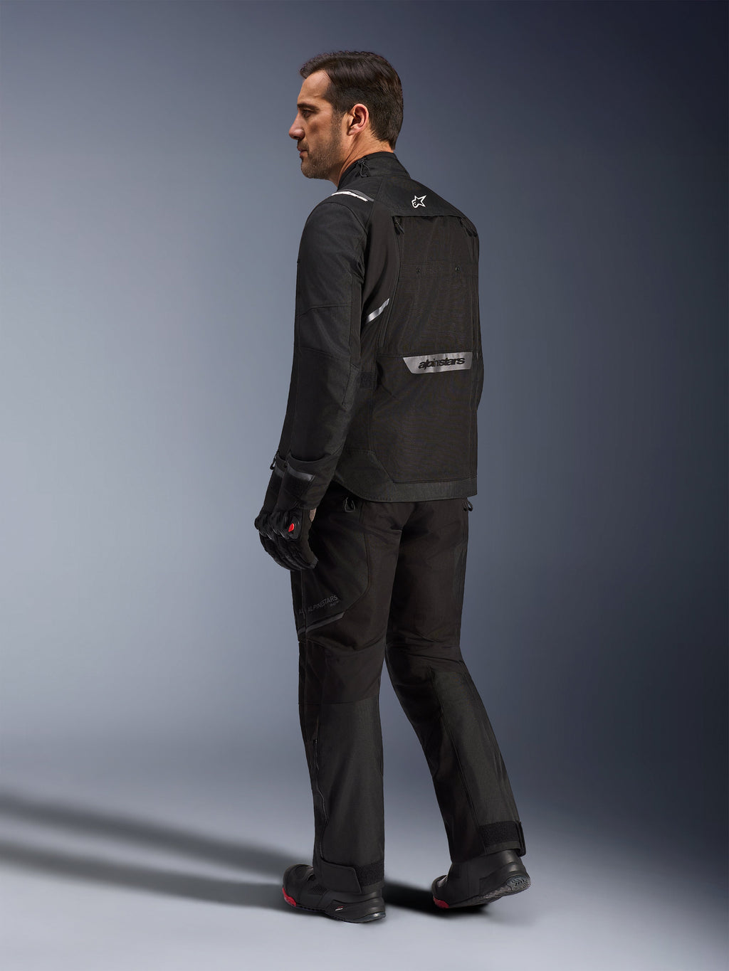 Alpinestars Cusco Drystar® Jacke, schwarze Motorradjacke, Rückansicht eines männlichen Models in kompletter Fahrausrüstung, reflektierendes Branding auf dem Rücken, Textilkonstruktion mit technischen Einsätzen, abgestimmtes Outfit für Road-Adventure-Touren