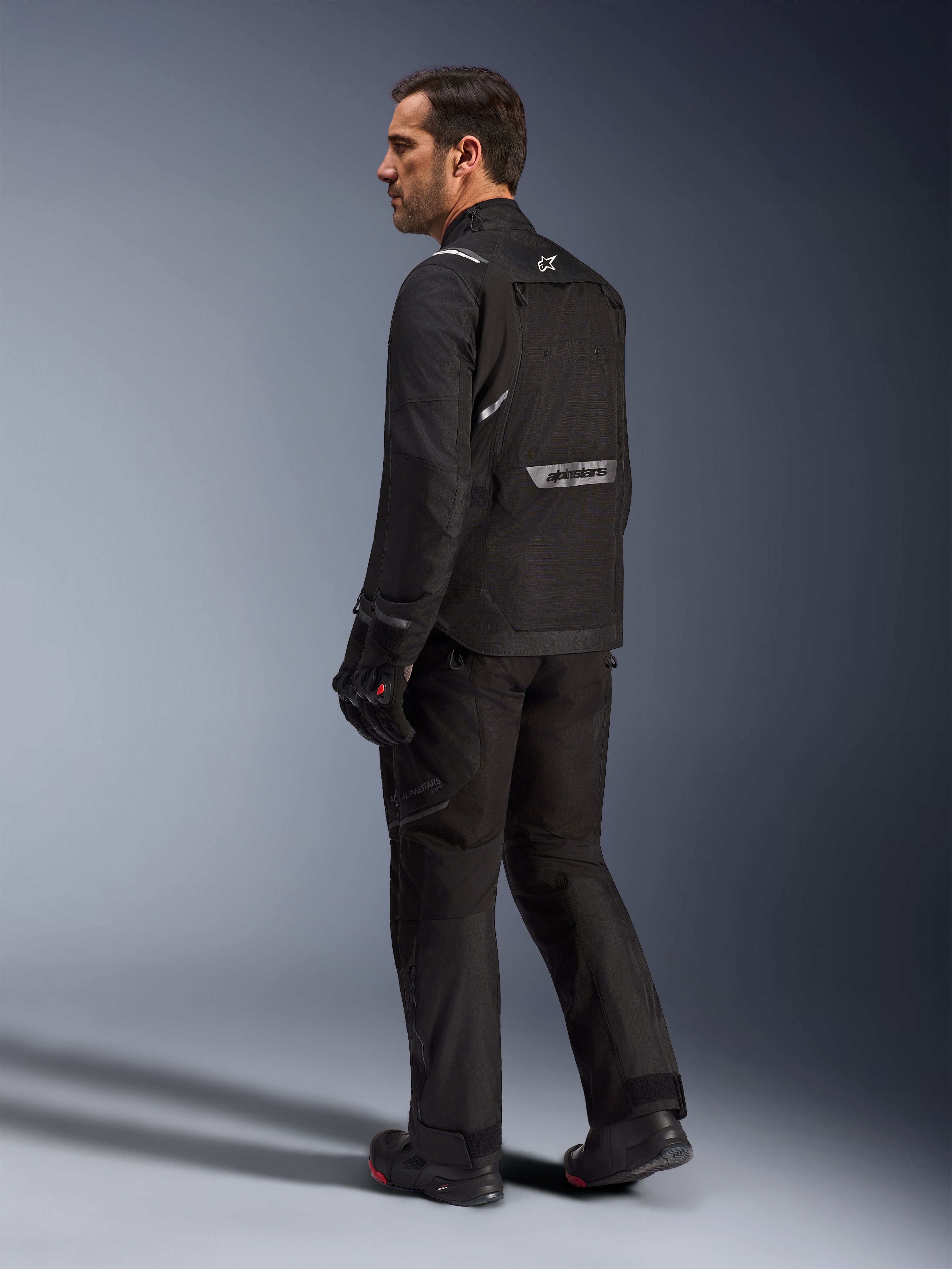 Alpinestars Cusco Drystar® Jacke, schwarze Motorradjacke, Rückansicht eines männlichen Models in kompletter Fahrausrüstung, reflektierendes Branding auf dem Rücken, Textilkonstruktion mit technischen Einsätzen, abgestimmtes Outfit für Road-Adventure-Touren