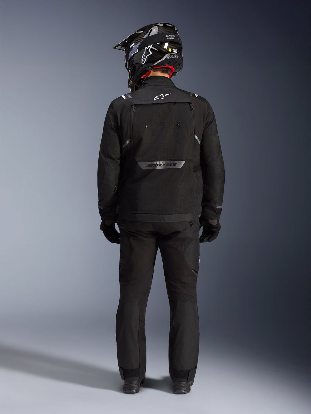 Alpinestars Cusco Drystar Jacke, Motorrad-Adventure-Jacke, Schwarz, Rückansicht getragen mit passender Hose und einem schwarz-weißen Helm, mit reflektierendem Alpinestars-Branding und großen Belüftungspanelen, strapazierfähige Textilkonstruktion für Adventure-Touren.