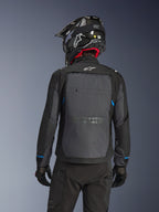 Rückansicht eines Fahrers in der Alpinestars Cusco Drystar® Jacke in Schwarz und Deep Gray, mit einem großen grauen Belüftungspaneel am Rücken und weißem Logo, kombiniert mit einem schwarzen Offroad-Helm und schwarzer Fahrausrüstung für Adventure Touring.