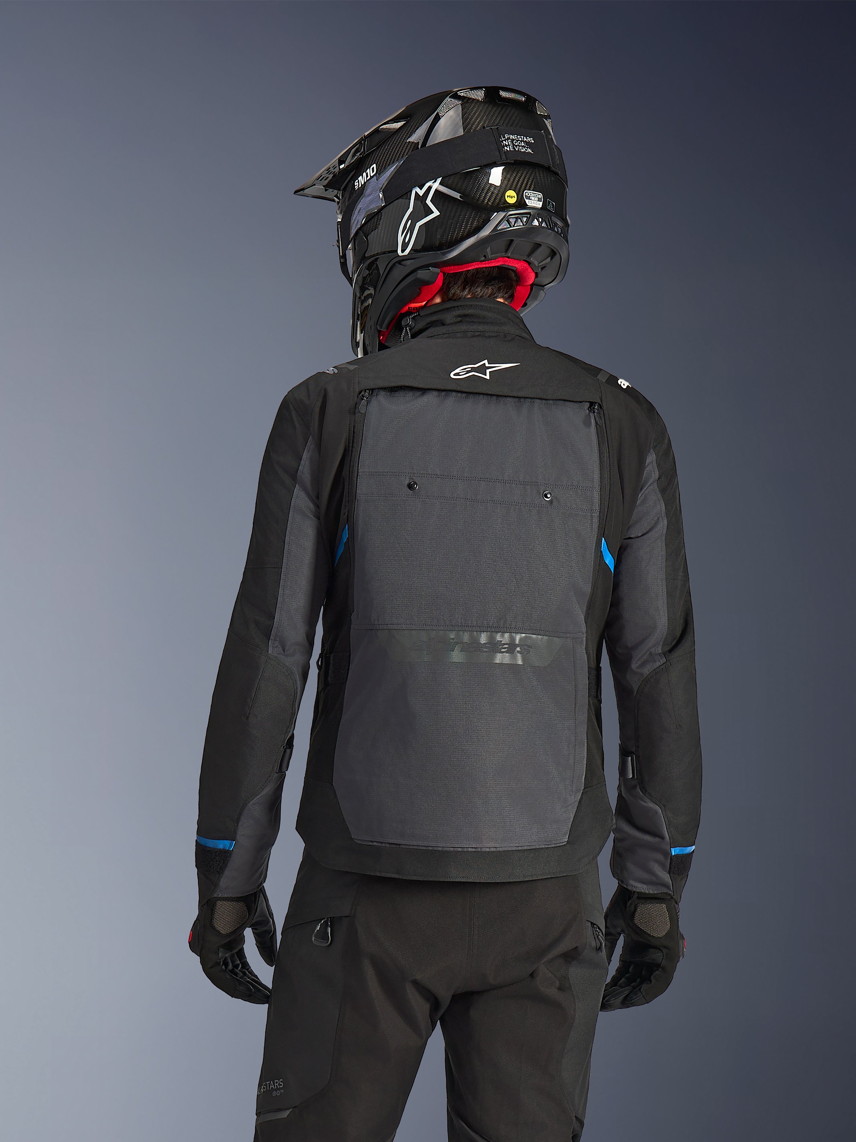 Rückansicht eines Fahrers in der Alpinestars Cusco Drystar® Jacke in Schwarz und Deep Gray, mit einem großen grauen Belüftungspaneel am Rücken und weißem Logo, kombiniert mit einem schwarzen Offroad-Helm und schwarzer Fahrausrüstung für Adventure Touring.