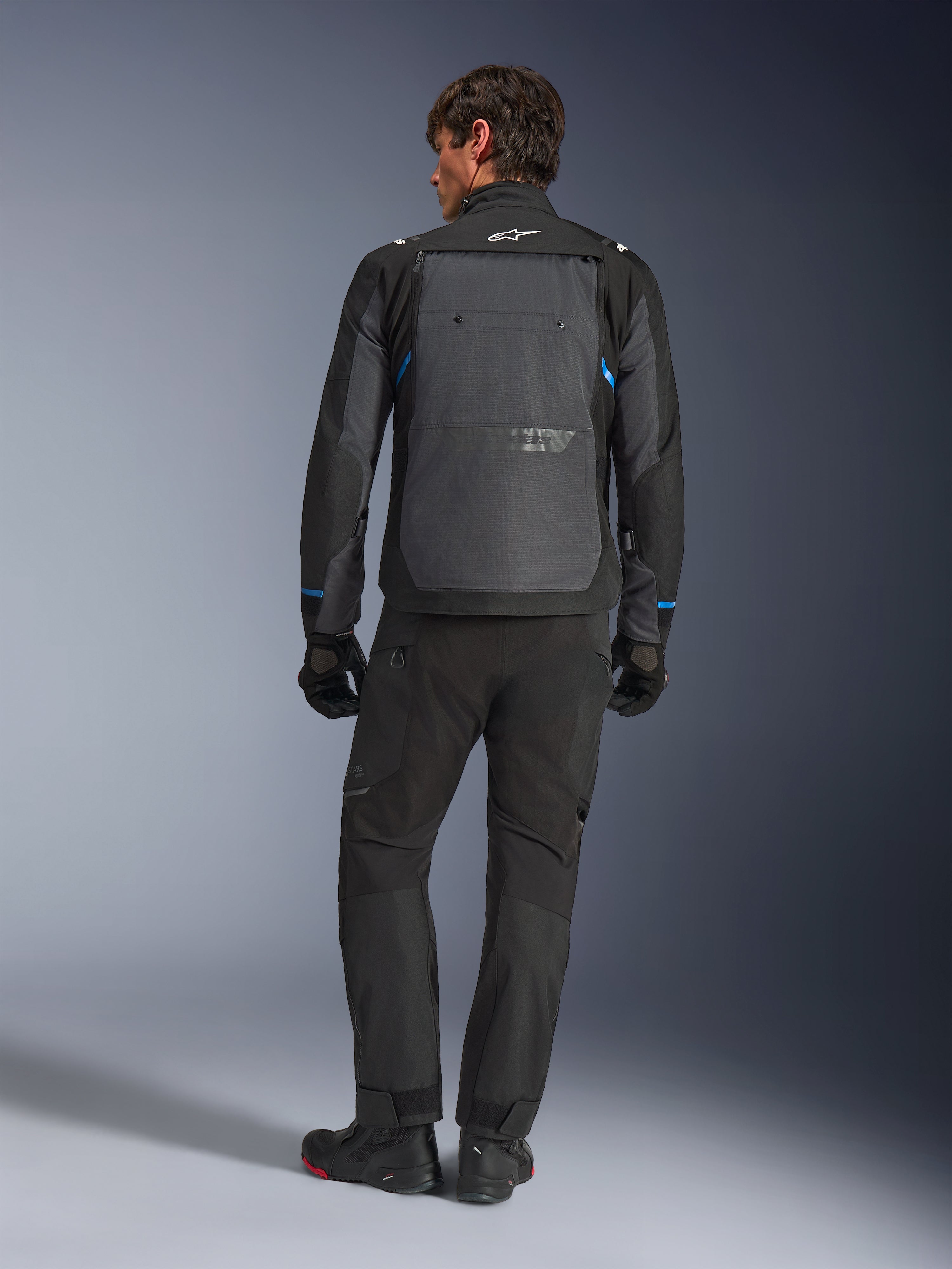 Alpinestars Cusco Drystar® Jacke, Schwarz und Dunkelgrau mit blauen Akzenten, Rückansicht, getragen mit passender schwarzer Adventure-Hose und Handschuhen, mit großem grauen Rückeneinsatz und weißem Logo am Kragen, konzipiert für Straßen- und Adventure-Touren