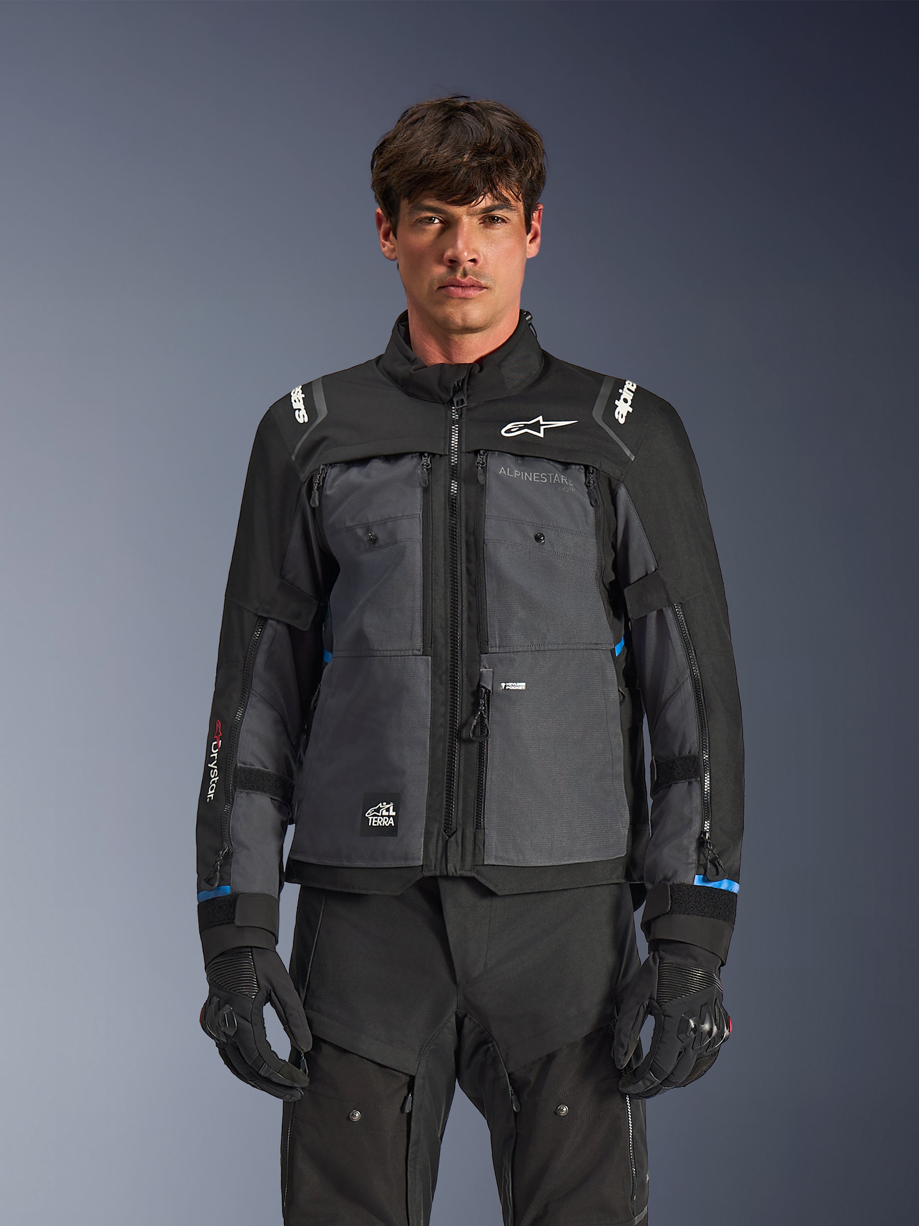 Alpinestars Cusco Drystar® Jacke, Motorrad-Tourenjacke, Schwarz und Dunkelgrau, Vorderansicht am Modell, mit Alpinestars-Logos auf Brust und Schultern, Drystar®-Branding am Ärmel, technisches Multi-Pocket-Design mit Belüftungsreißverschlüssen, kombiniert mit schwarzer Motorradausrüstung