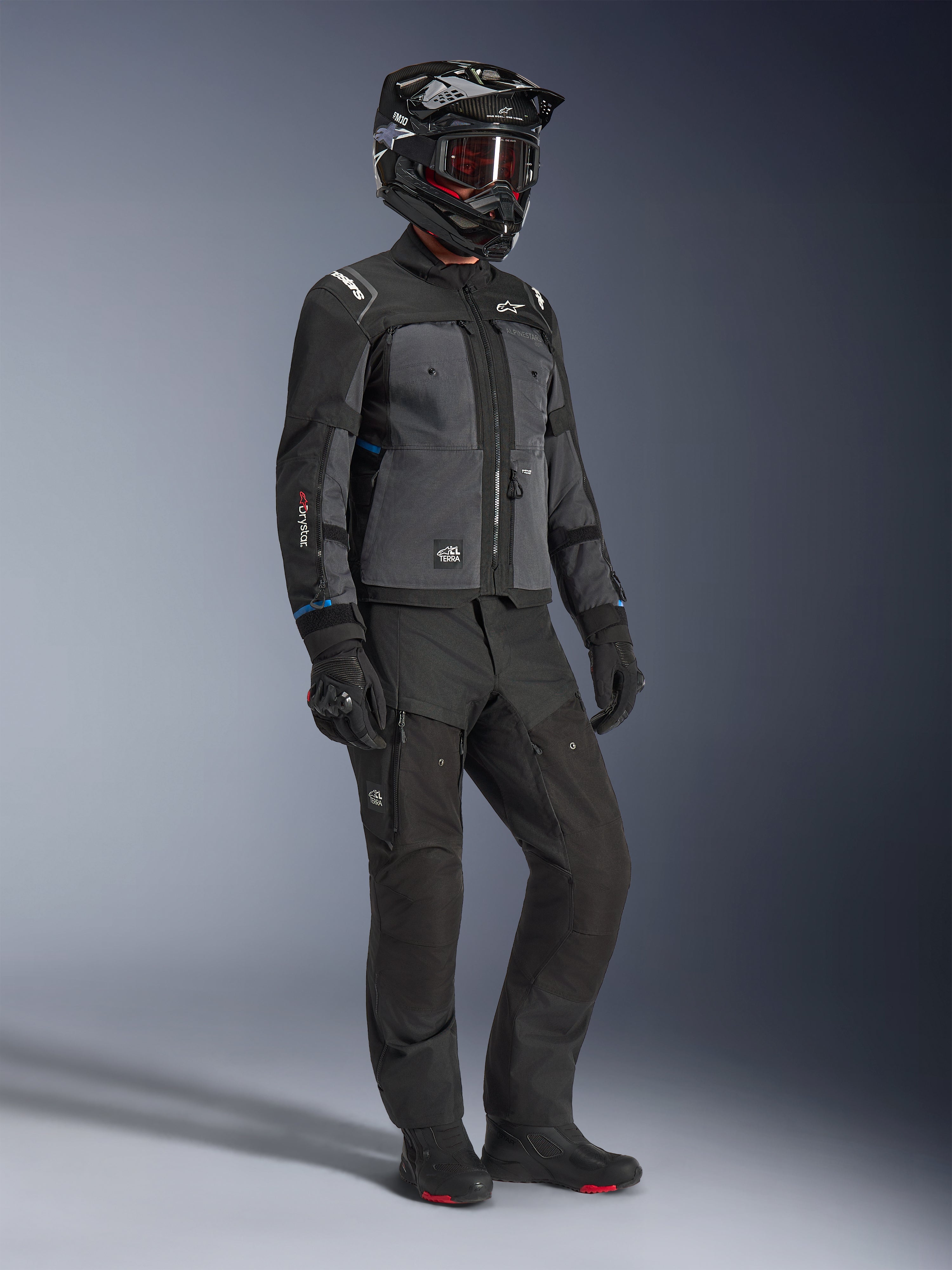 Model trägt die Alpinestars Cusco Drystar® Jacke in Schwarz und Deep Gray, kombiniert mit kompletter Motorrad-Ausrüstung inklusive schwarzem Helm, passender technischer Hose, Handschuhen und Stiefeln, mit Ripstop- und Softshell-Konstruktion für Road- und Adventure-Touren
