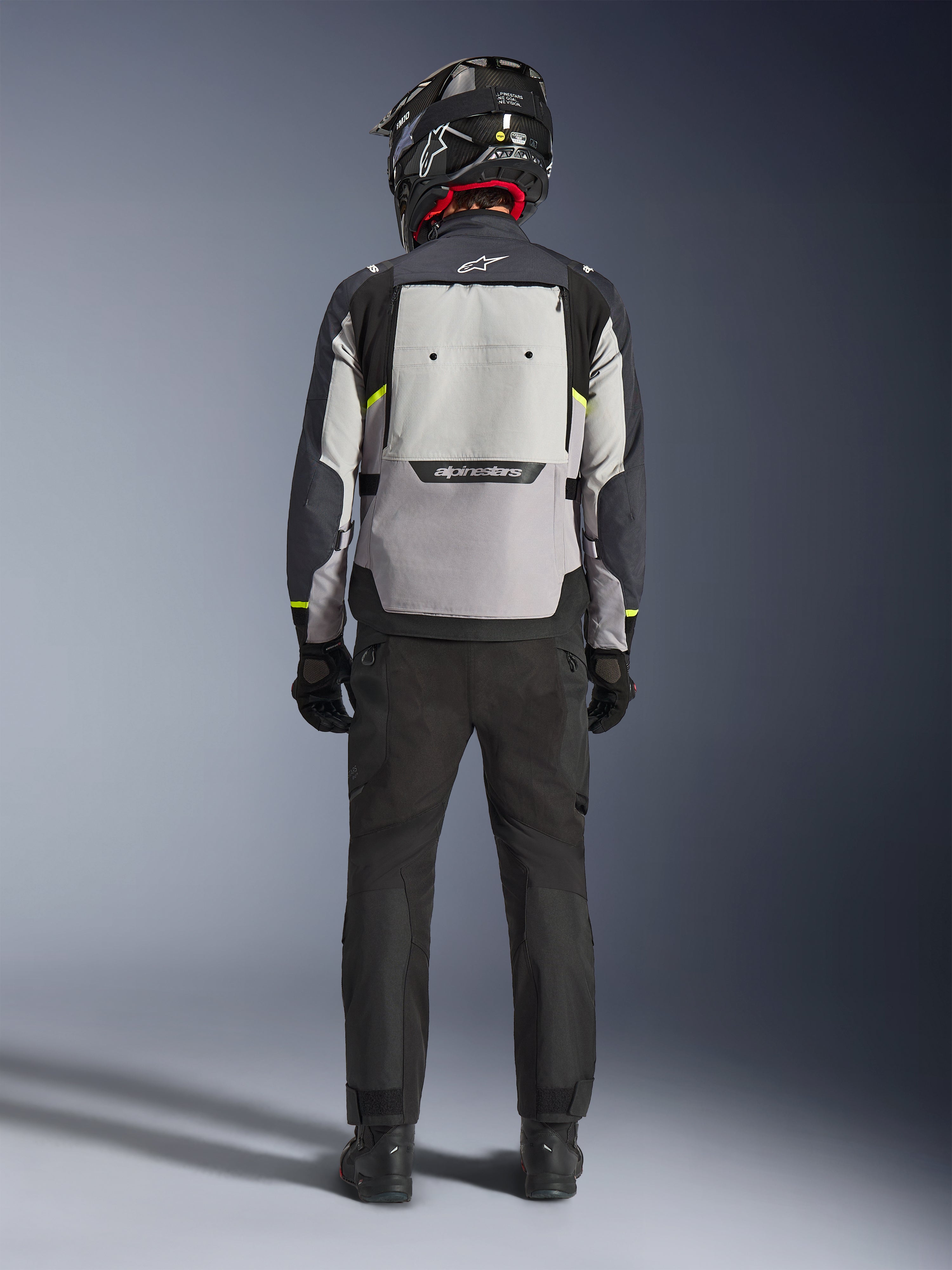 Motorradfahrer in der Rückansicht mit der Alpinestars Cusco Drystar® Jacke in Ice Gray, Mid Gray und Deep Gray, mit großem Belüftungspanel am Rücken und Alpinestars-Branding, kombiniert mit einer schwarzen Motorradhose und einem schwarzen Helm, Adventure-Touring-Motorradbekleidung.