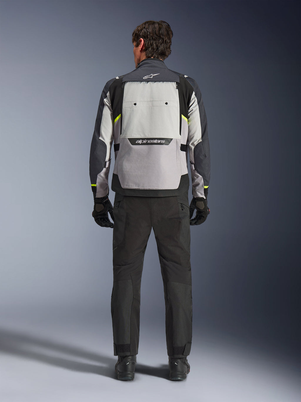 Rückansicht eines Fahrers, der die Alpinestars Cusco Drystar® Jacke in Ice Gray, Mid Gray und Deep Gray trägt, kombiniert mit einer schwarzen Motorradhose und Handschuhen, wobei das große Belüftungspanel am Rücken und das Alpinestars-Branding hervorgehoben werden.
