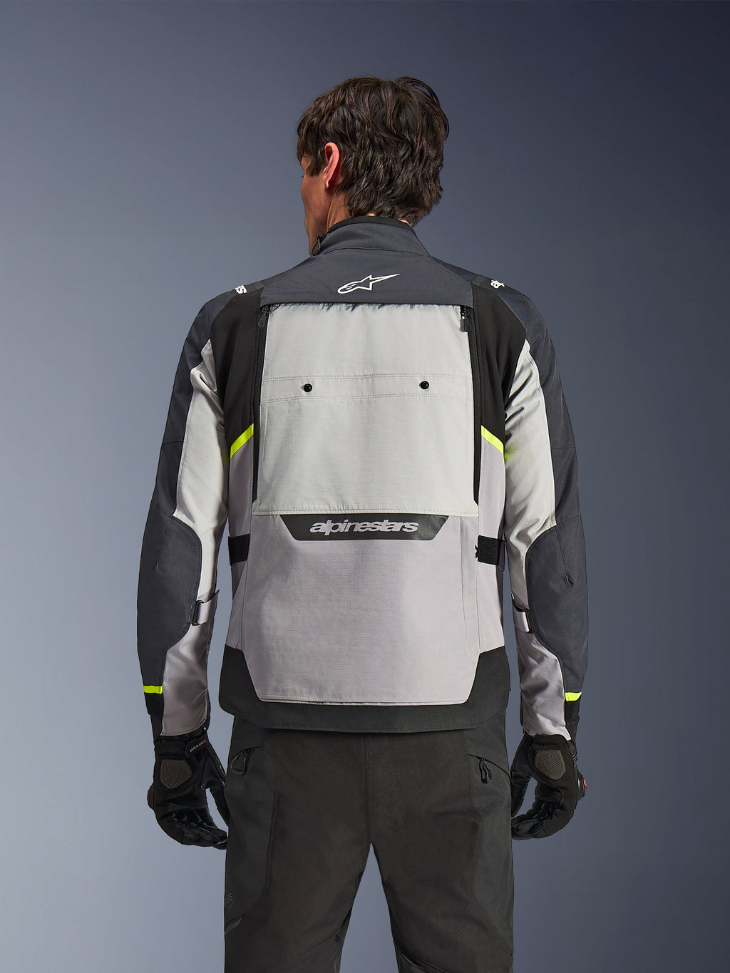 Rückansicht eines Fahrers in der Alpinestars Cusco Drystar® Jacke in Ice Gray, Mid Gray und Deep Gray, mit neongelben Akzenten, einem großen Belüftungspanel am Rücken und markantem Alpinestars-Branding, kombiniert mit schwarzer Fahrhose und Handschuhen.