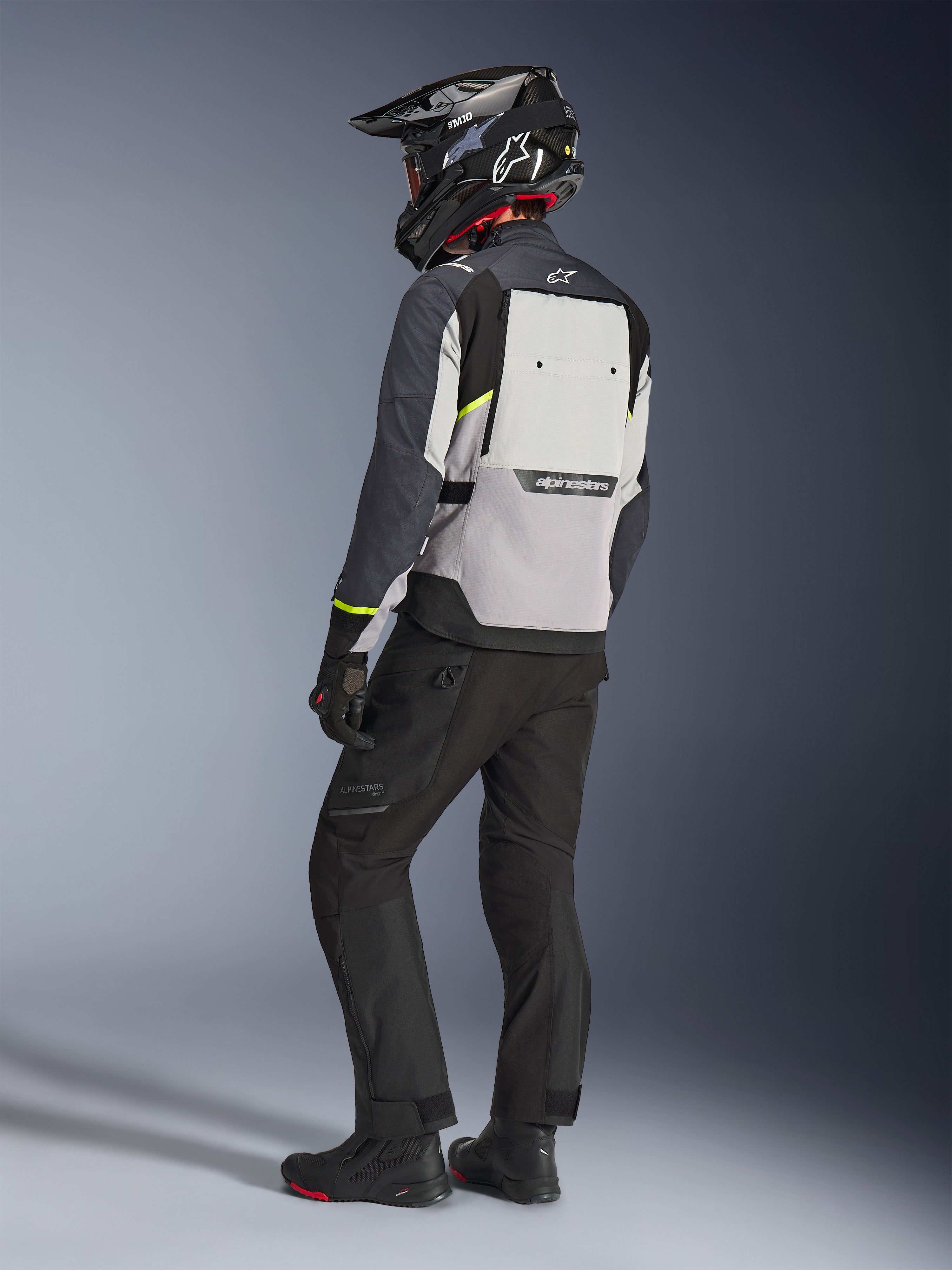 Alpinestars Cusco Drystar® Jacke in Ice Gray, Mid Gray und Deep Gray, Rückansicht an einem Fahrer. Ausgestattet mit neongelben Akzenten, einem großen Belüftungspanel am Rücken mit Branding und einer strapazierfähigen Textilkonstruktion. Getragen mit einer schwarzen Motorradhose und einem Adventure-Helm für einen kompletten Touring-Look.