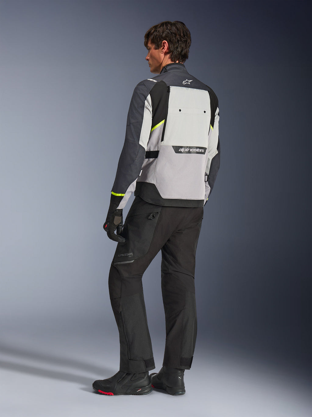 Alpinestars Cusco Drystar® Jacke in Ice Gray, Mid Gray und Deep Gray, Rückansicht am Modell, Adventure-Tourenjacke mit neongelben Akzenten, getragen mit schwarzer Motorradhose und Stiefeln, mit großem Belüftungspaneel am Rücken und Alpinestars-Branding.