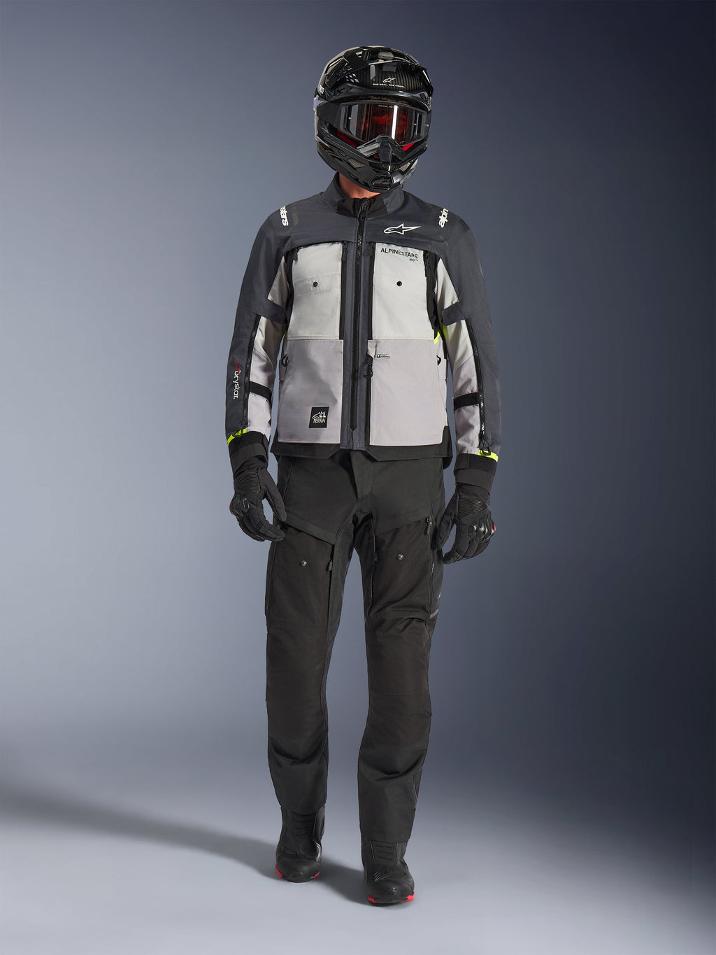 Alpinestars Cusco Drystar® Jacke, Ice Gray Mid Gray Deep Gray, Adventure-Motorradjacke kombiniert mit schwarzer Motorradhose, schwarzen Handschuhen und einem schwarzen Helm mit Brille, mit farblich abgesetzten grauen Einsätzen und technischen Details für Straßen- und Adventure-Touren.
