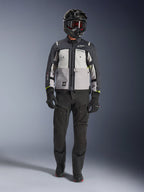 Alpinestars Cusco Drystar® Jacke, Ice Gray Mid Gray Deep Gray, Adventure-Motorradjacke kombiniert mit schwarzer Motorradhose, schwarzen Handschuhen und einem schwarzen Helm mit Brille, mit farblich abgesetzten grauen Einsätzen und technischen Details für Straßen- und Adventure-Touren.
