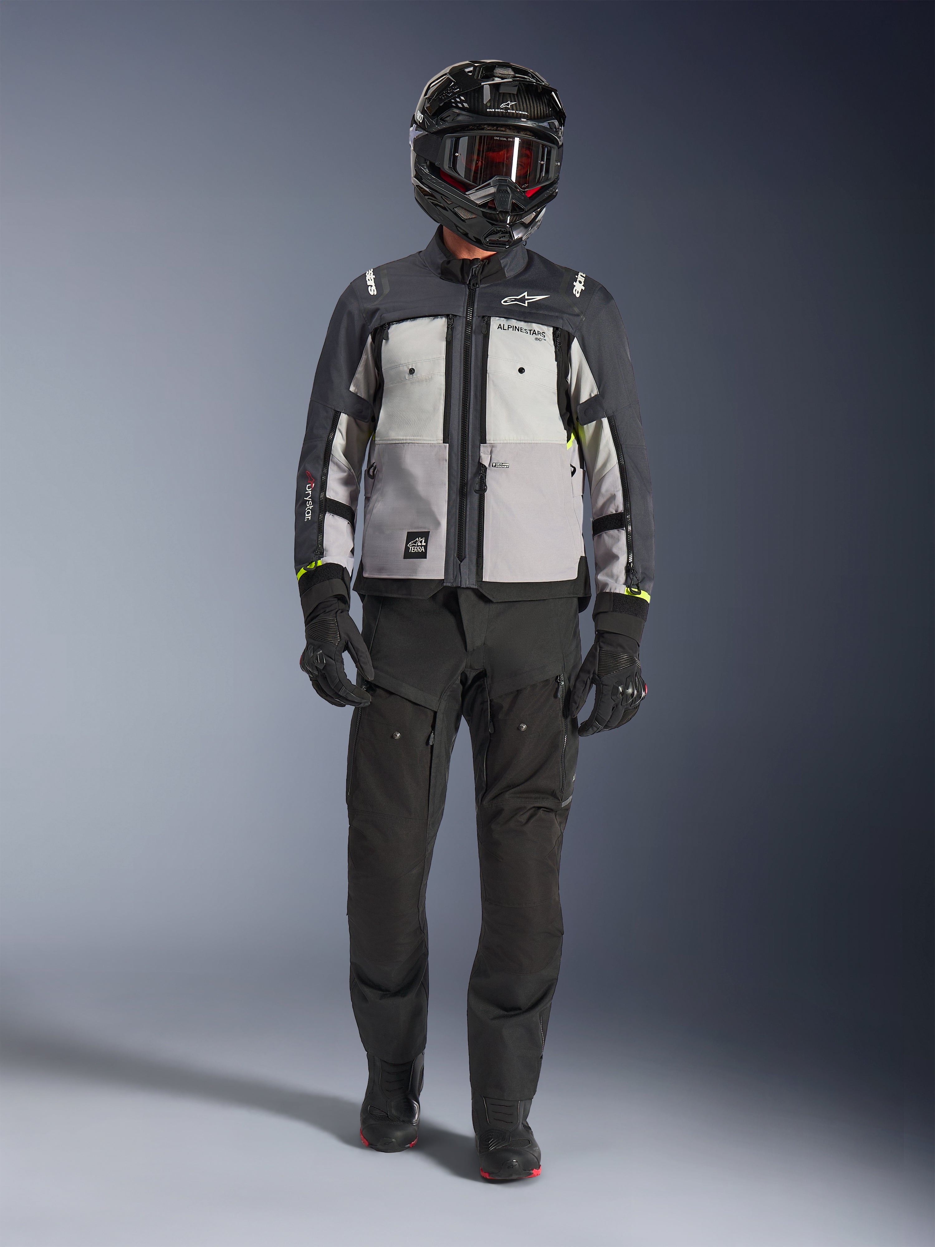 Alpinestars Cusco Drystar® Jacke, Ice Gray Mid Gray Deep Gray, Adventure-Motorradjacke kombiniert mit schwarzer Motorradhose, schwarzen Handschuhen und einem schwarzen Helm mit Brille, mit farblich abgesetzten grauen Einsätzen und technischen Details für Straßen- und Adventure-Touren.