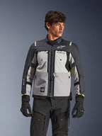 Alpinestars Cusco Drystar® Jacke, Adventure-Touren-Motorradjacke, Ice Gray, Mid Gray und Deep Gray, getragen von einem Modell mit schwarzer Hose und Handschuhen, mit einem mehrteiligen technischen Textildesign, Brusttaschen, 'Drystar'-Branding auf dem Ärmel und dezenten neongelben Akzenten an den Ärmelbündchen.