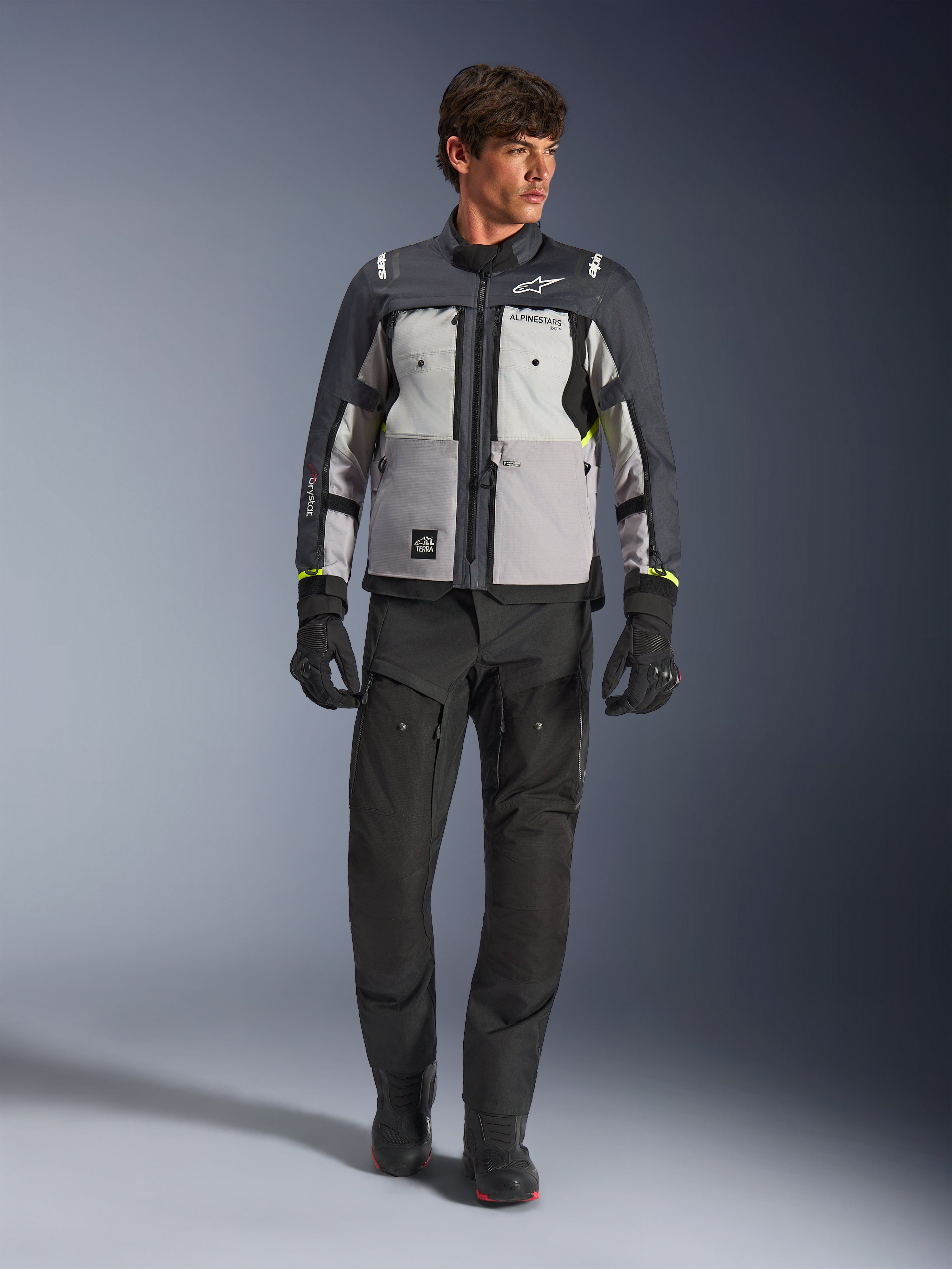 Alpinestars Cusco Drystar® Jacke in Ice Gray, Mid Gray und Deep Gray, getragen mit schwarzer Adventure-Motorradhose und Handschuhen, Textil-Adventure-Tourenjacke mit mehrfarbigen grauen Einsätzen und neongelben Akzenten, Studioaufnahme eines männlichen Models.