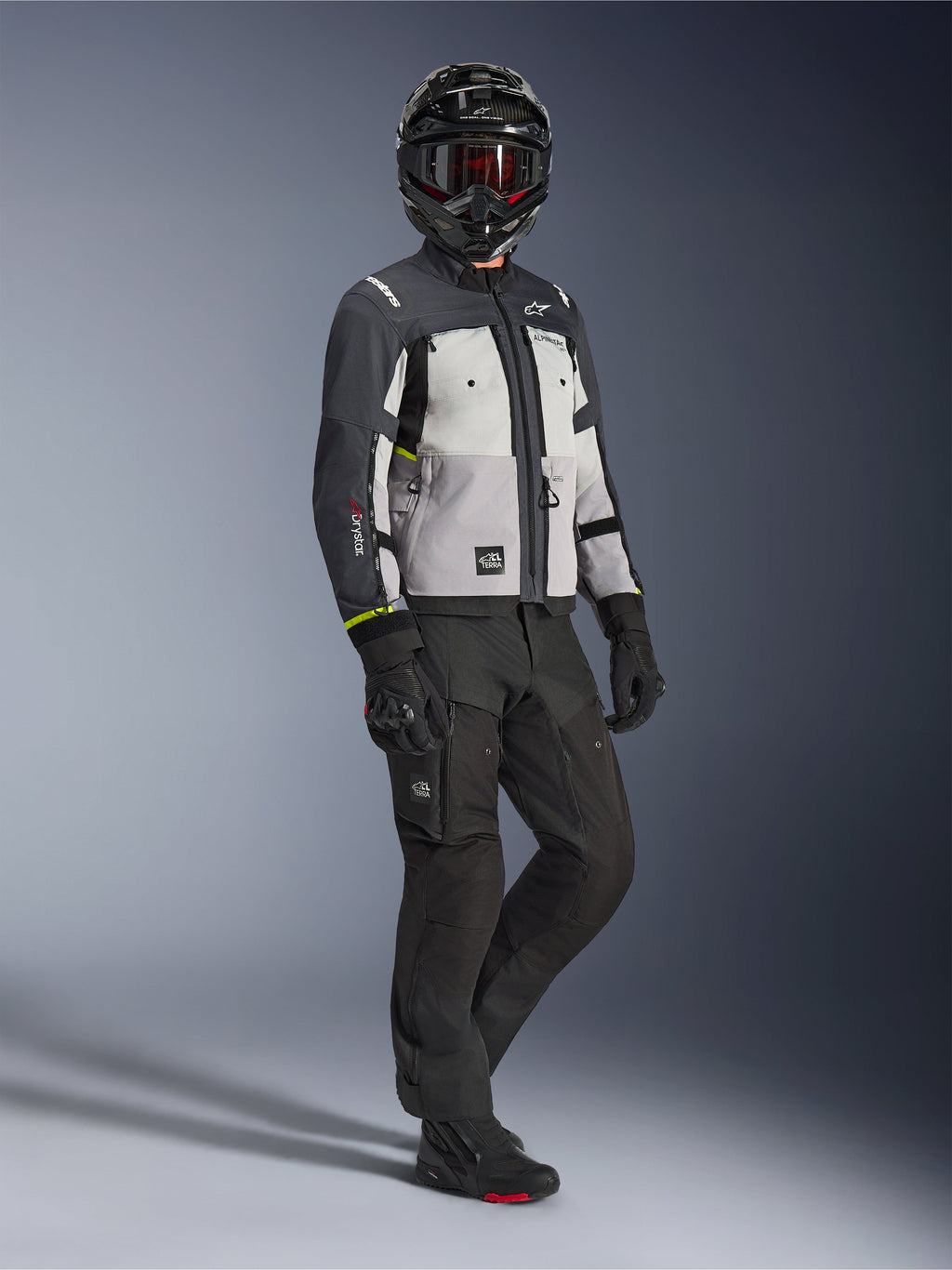 Alpinestars Cusco Drystar® Jacke in Ice Gray Mid Gray Deep Gray, Ganzkörperaufnahme eines Fahrers in kompletter Adventure-Ausrüstung, einschließlich eines schwarzen Helms mit roter Brille, passender schwarzer Hose und Handschuhen, Textil-Motorradjacke mit direkten Belüftungspaneelen und wasserdichter Membran