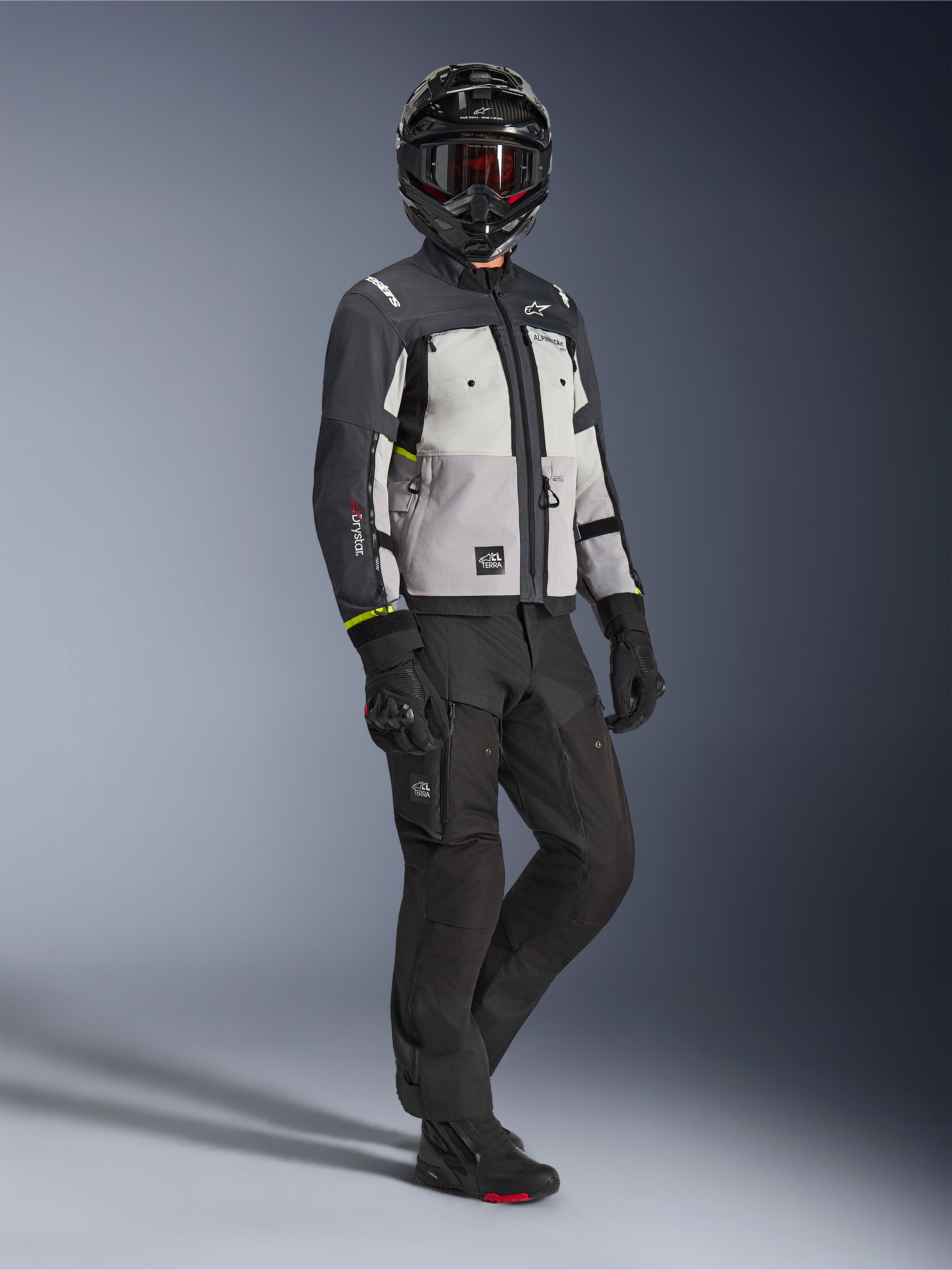 Alpinestars Cusco Drystar® Jacke in Ice Gray Mid Gray Deep Gray, Ganzkörperaufnahme eines Fahrers in kompletter Adventure-Ausrüstung, einschließlich eines schwarzen Helms mit roter Brille, passender schwarzer Hose und Handschuhen, Textil-Motorradjacke mit direkten Belüftungspaneelen und wasserdichter Membran