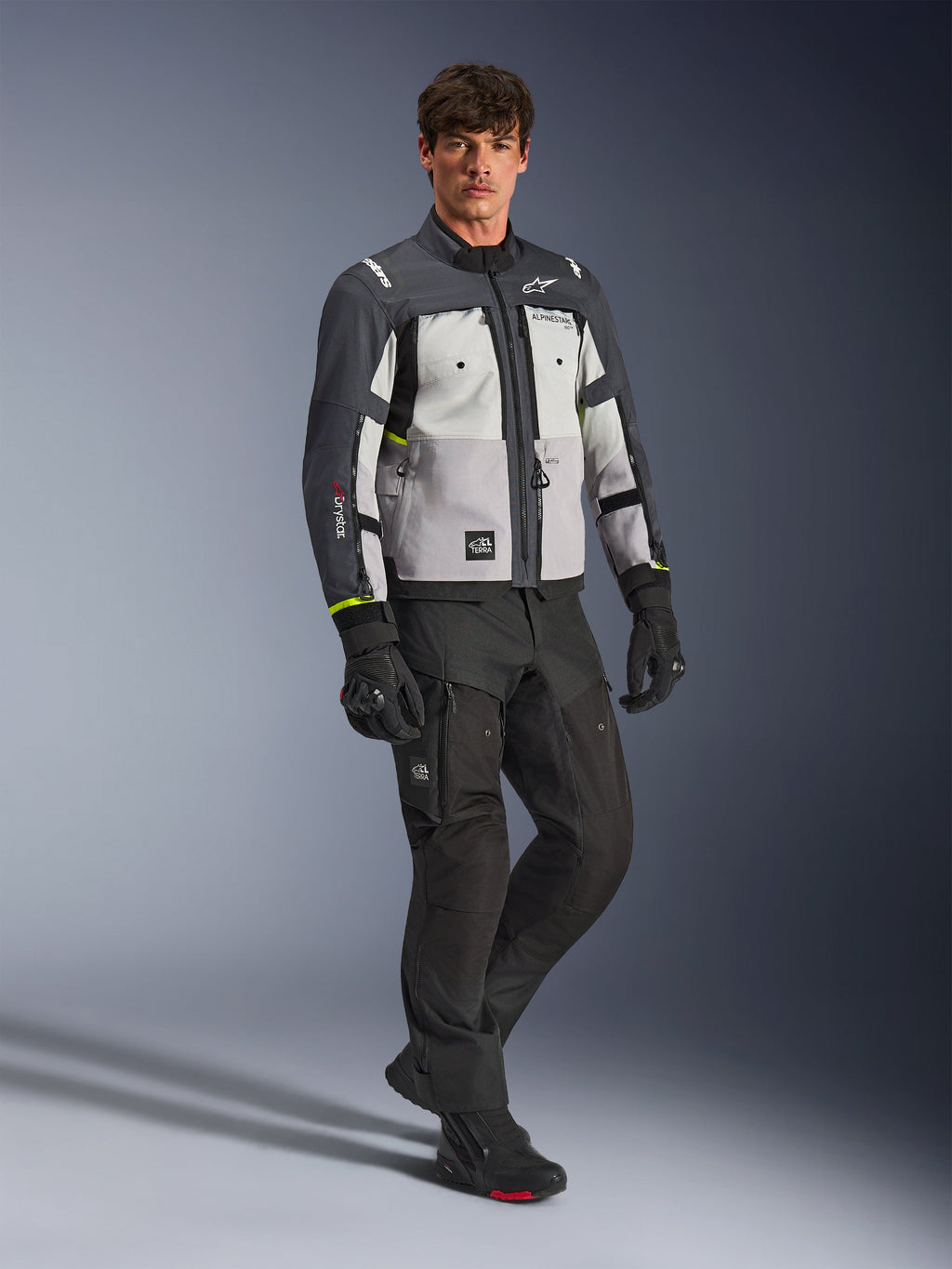 Alpinestars Cusco Drystar® Jacke, Motorrad-Adventure-Jacke, Eisgrau, Mittelgrau und Dunkelgrau, getragen von einem Model mit schwarzer Hose und Handschuhen, mehrfarbige graue Einsätze mit strapazierfähiger Textilkonstruktion, mit Belüftungsreißverschlüssen und Alpinestars-Branding, professionelle Studiofotografie für Straßen- und Adventure-Fahrbekleidung