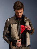 Alpinestars Cusco Drystar® Jacke, Walnut Aluminum Black, getragen von einem Mann, der die Brustprotektorentasche mit einem roten Protektoreinsatz zeigt; Textil-Motorradjacke mit Belüftungsöffnungen mit Reißverschluss und Branding für Adventure-Touring.