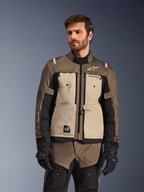 Alpinestars Cusco Drystar® Jacke, Adventure-Motorradjacke, Walnut Aluminum Black, von einem Modell mit schwarzen Handschuhen getragen, mit hellbraunen Brustpaneelen und schwarzen Ärmeln mit Alpinestars-Branding und All-Terra-Patch, schützende Allwetter-Tourenbekleidung