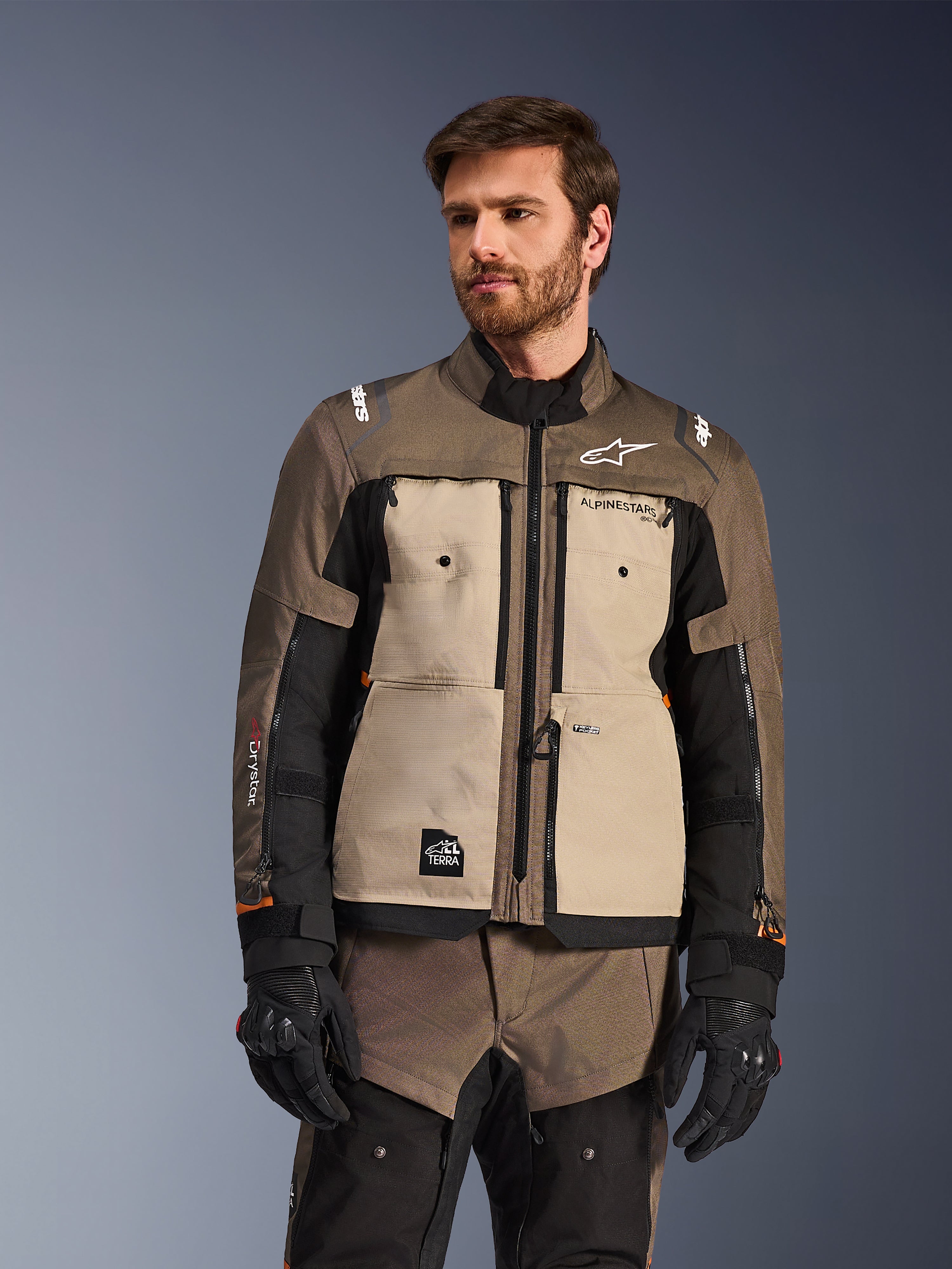 Alpinestars Cusco Drystar® Jacke, Adventure-Motorradjacke, Walnut Aluminum Black, von einem Modell mit schwarzen Handschuhen getragen, mit hellbraunen Brustpaneelen und schwarzen Ärmeln mit Alpinestars-Branding und All-Terra-Patch, schützende Allwetter-Tourenbekleidung