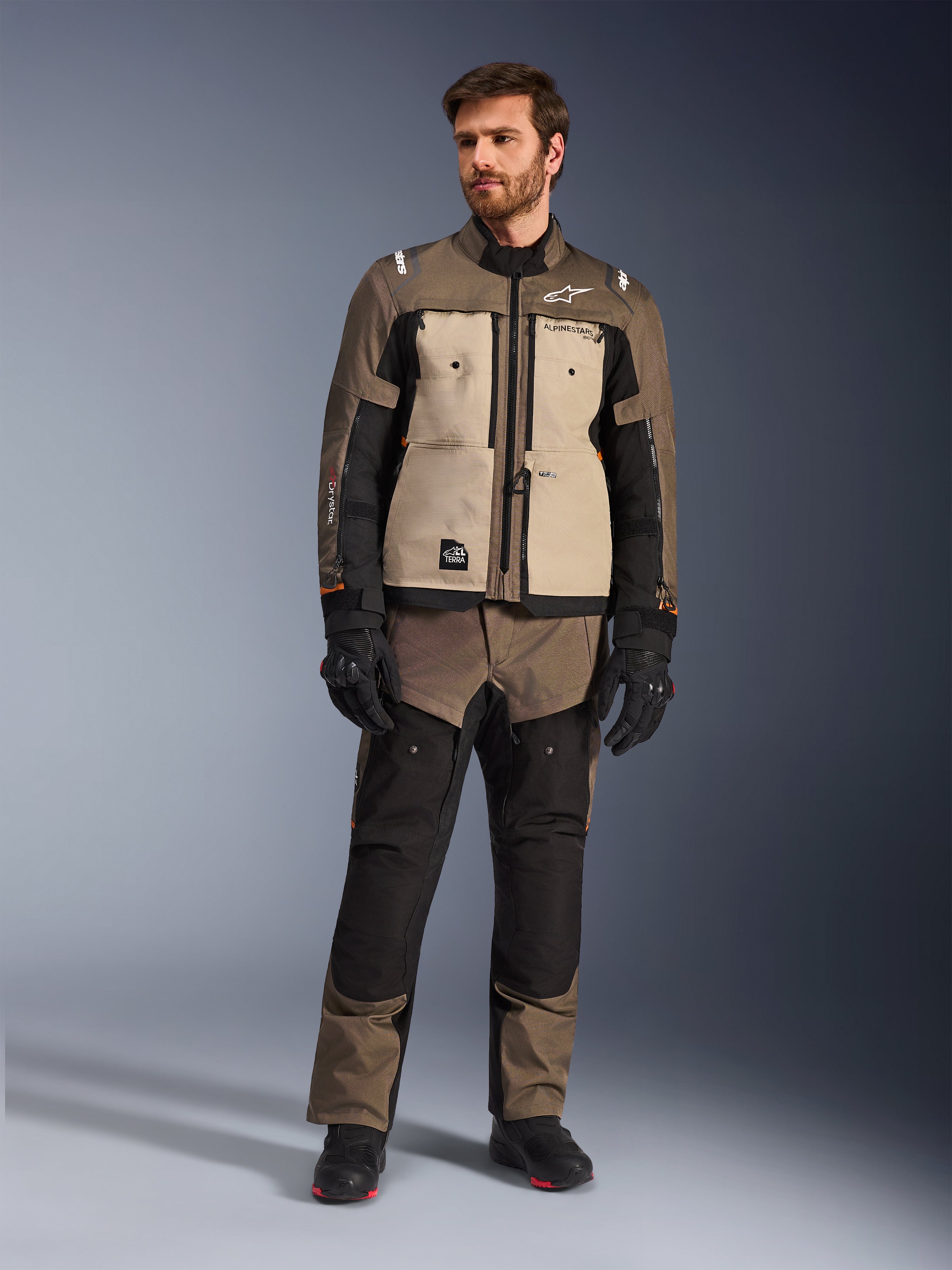 Alpinestars Cusco Drystar® Jacke in Walnut Aluminum Black, getragen von einem männlichen Fahrer mit passender Adventure-Tourenhose, schwarzen Handschuhen und Stiefeln, mit beigen Brustpaneelen, schwarzen Ärmeln mit dezenten orangefarbenen Akzenten und funktionellen Belüftungsreißverschlüssen für Motorrad-Straßenfahrten.