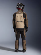 Fahrer in der Rückansicht mit der Alpinestars Cusco Drystar® Jacke in Walnut Aluminum Black, große aluminiumfarbene Rückentasche und orangefarbene Akzente, passende Adventure-Hose, schwarzer Offroad-Helm und Handschuhe, strapazierfähige Textil-Motorradausrüstung für Touren