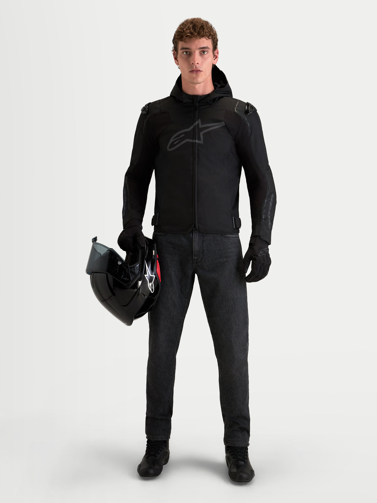 Alpinestars Zaca V2 Waterproof Jacket, Men's Black motorcycle textile jacket with hood and shoulder armor, worn by a male rider holding a helmet.