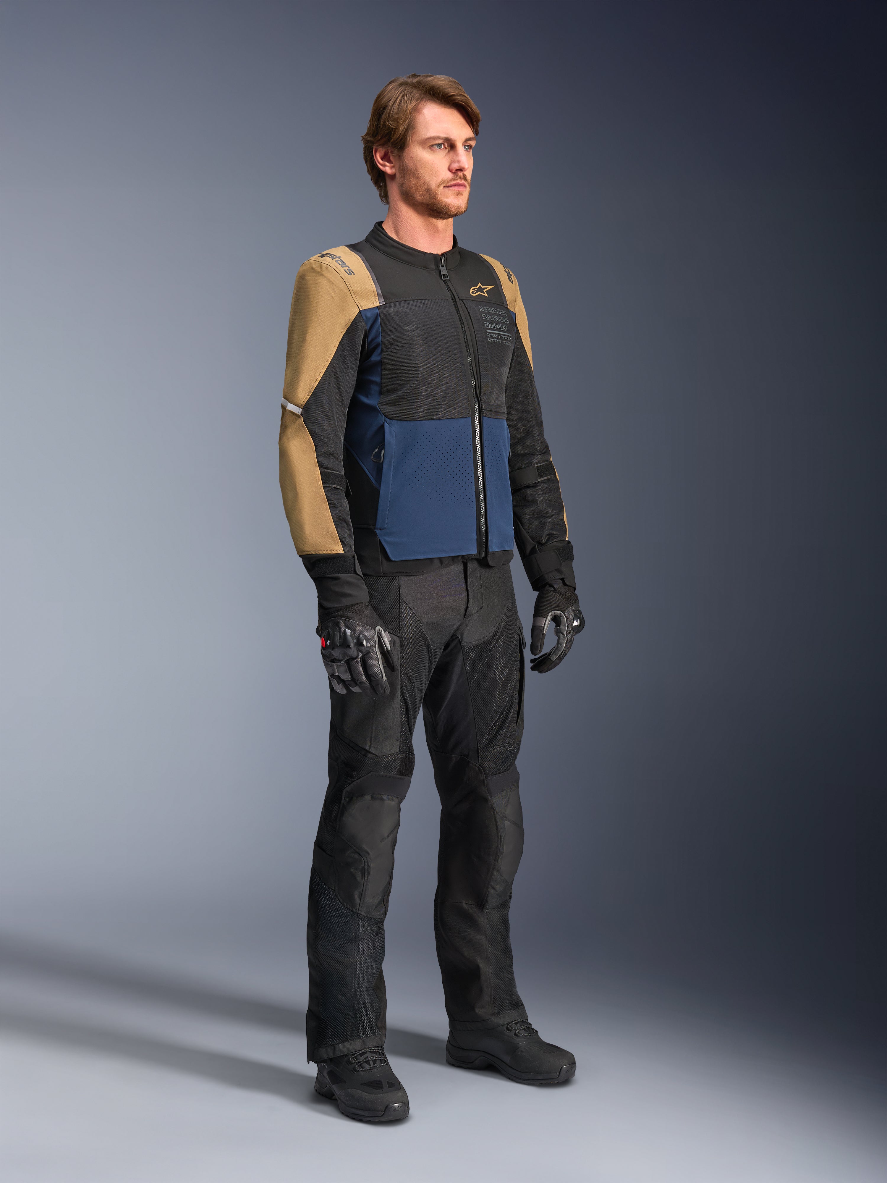 Männlicher Fahrer in der Alpinestars ST-2 Air Jacke in Deep Blue, Dark Khaki und Schwarz, mit großen Mesh-Einsätzen und Laserperforationen, kombiniert mit schwarzer Motorradhose, Handschuhen und Stiefeln für einen kompletten Sommer-Touren-Look.