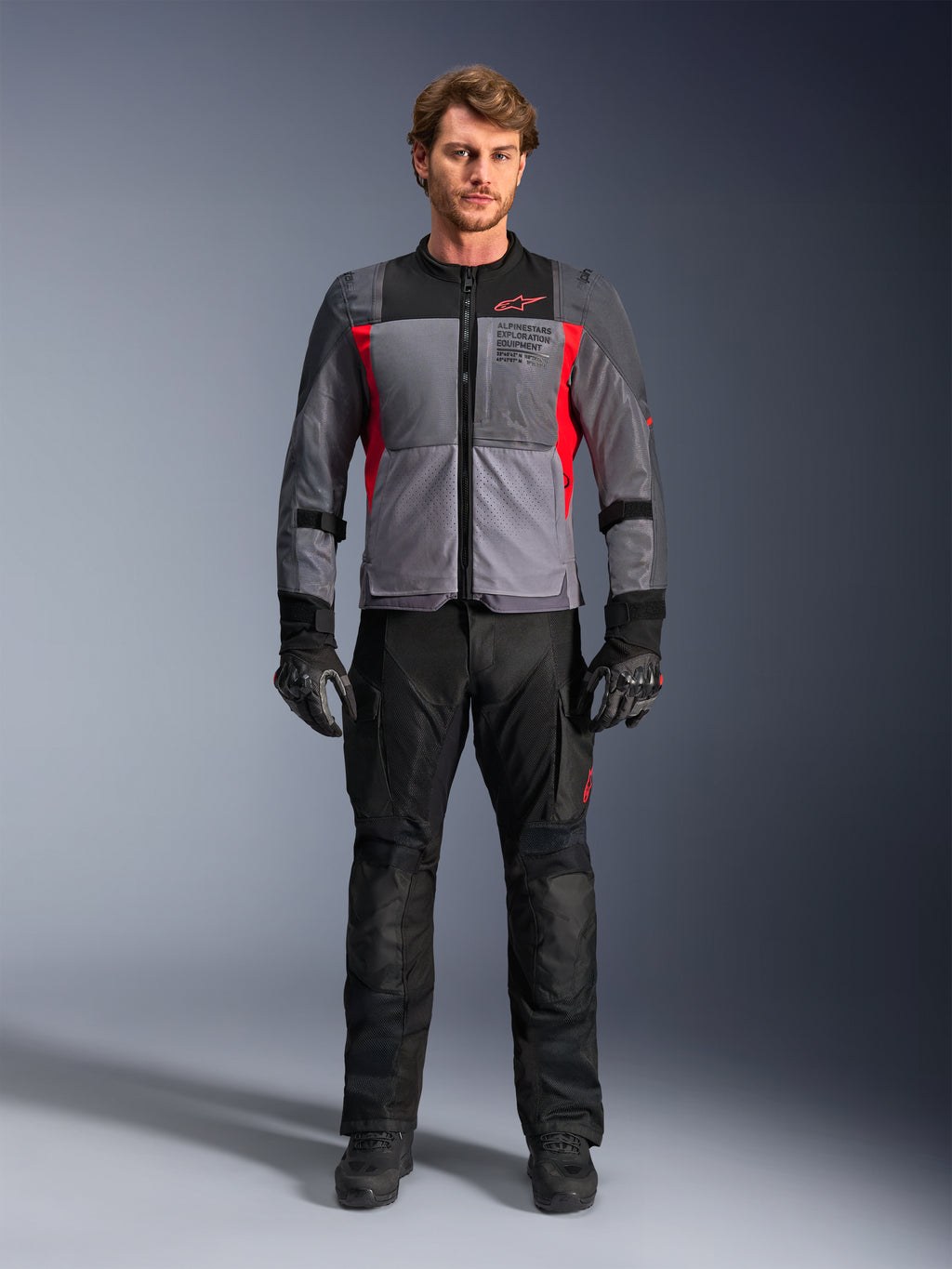 Alpinestars ST-2 Air Jacke in Deep Gray und Bright Red, getragen von einem männlichen Model mit schwarzer Motorradhose und Handschuhen, mit großen Mesh-Einsätzen für Luftzirkulation und modernem Exploration Equipment Branding