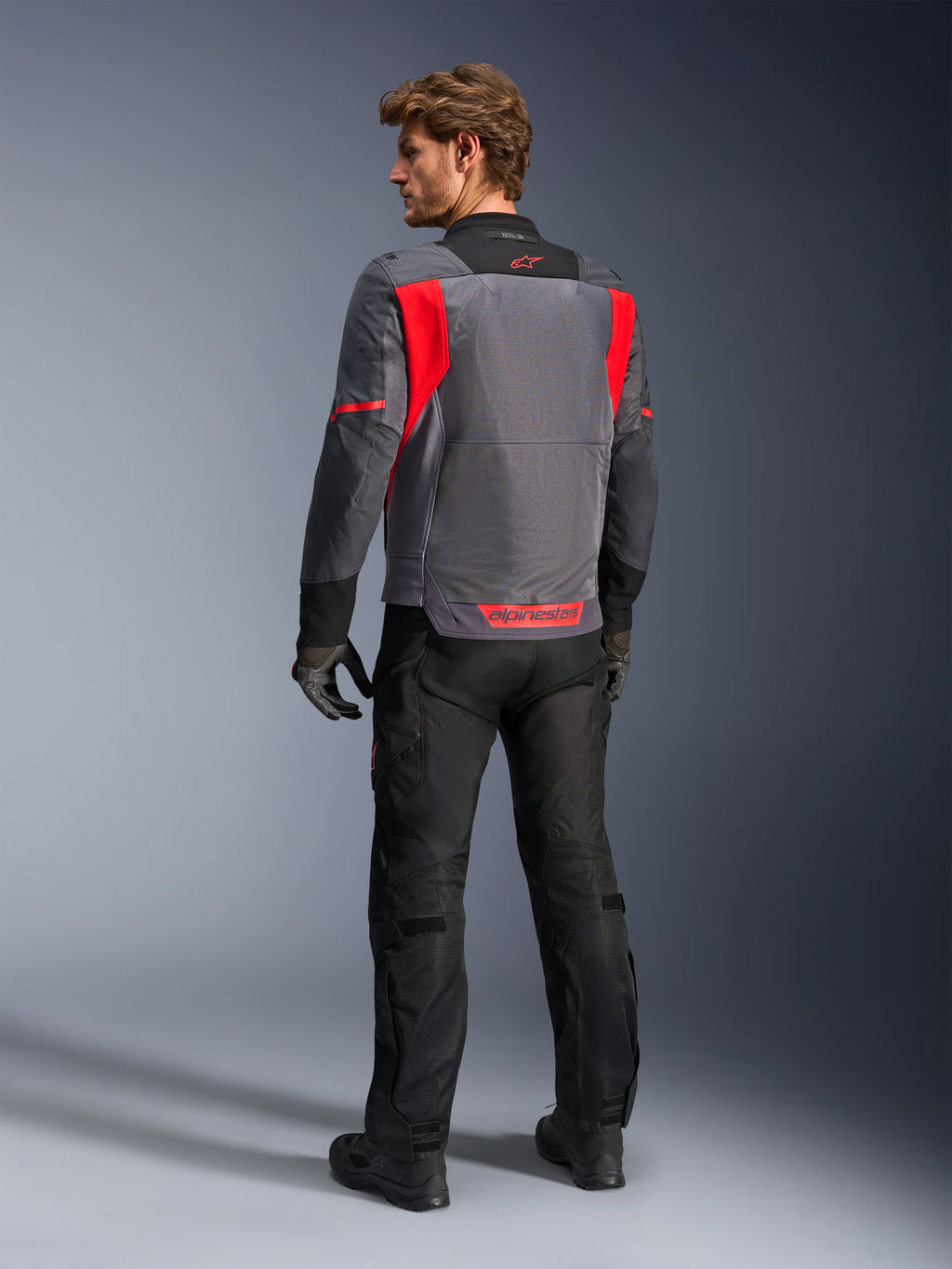 Männlicher Fahrer von hinten in der Alpinestars St-2 Air Jacke in Deep Gray, Dark Gray und Bright Red, präsentiert das große Mesh-Belüftungspanel am Rücken und das rote Branding, kombiniert mit einer schwarzen Motorradhose und grauen Handschuhen.