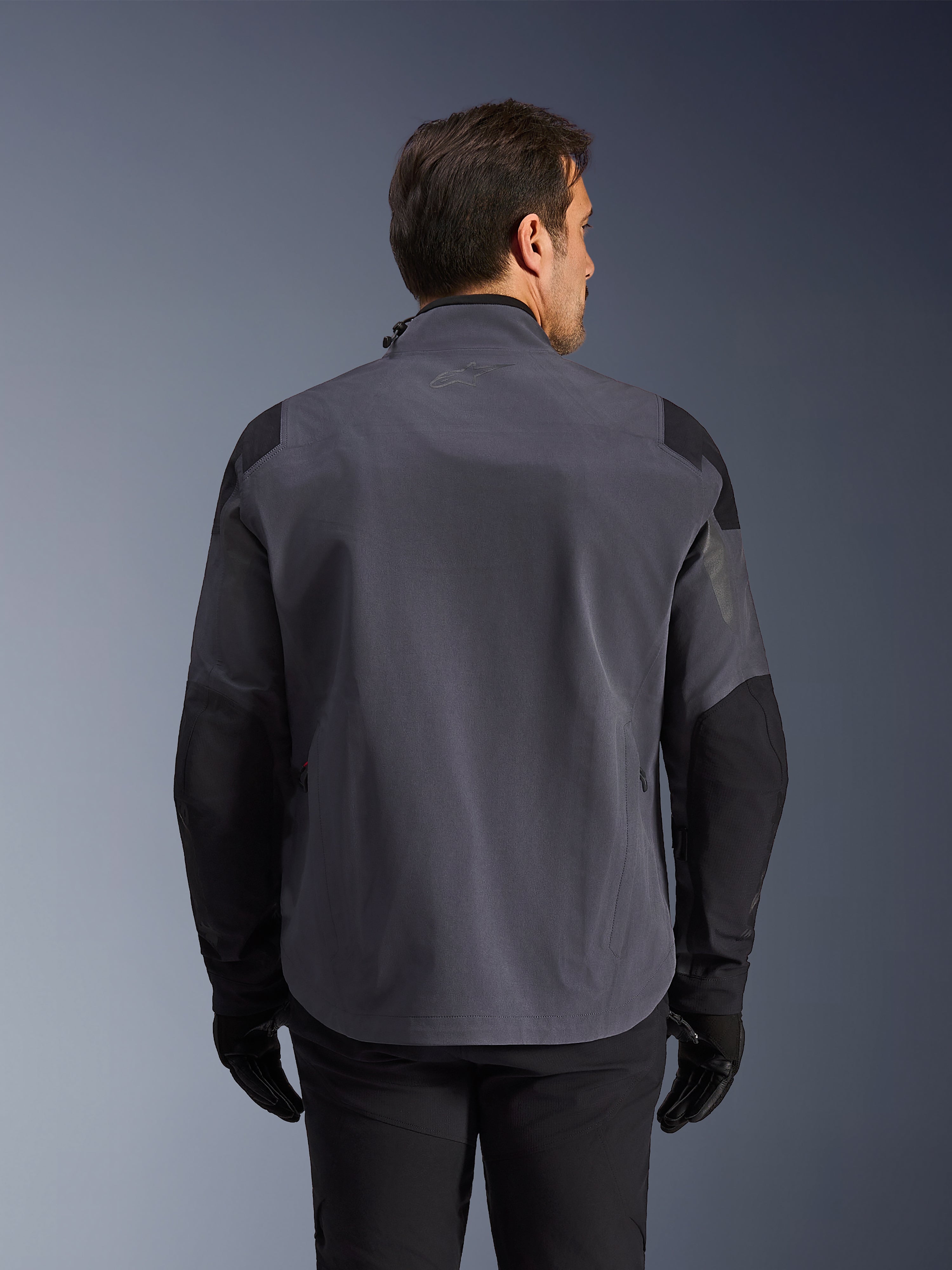 Alpinestars Maxdura Drystar®XF Laminierte Gepanzerte Jacke, technische Adventure-Jacke, Deep Gray und Schwarz, Rückansicht mit verstärkten Schulter- und Ellbogenpaneelen, dezentes Logo am oberen Rücken, getragen mit schwarzen Handschuhen und Hosen, entwickelt für Allwetterschutz.