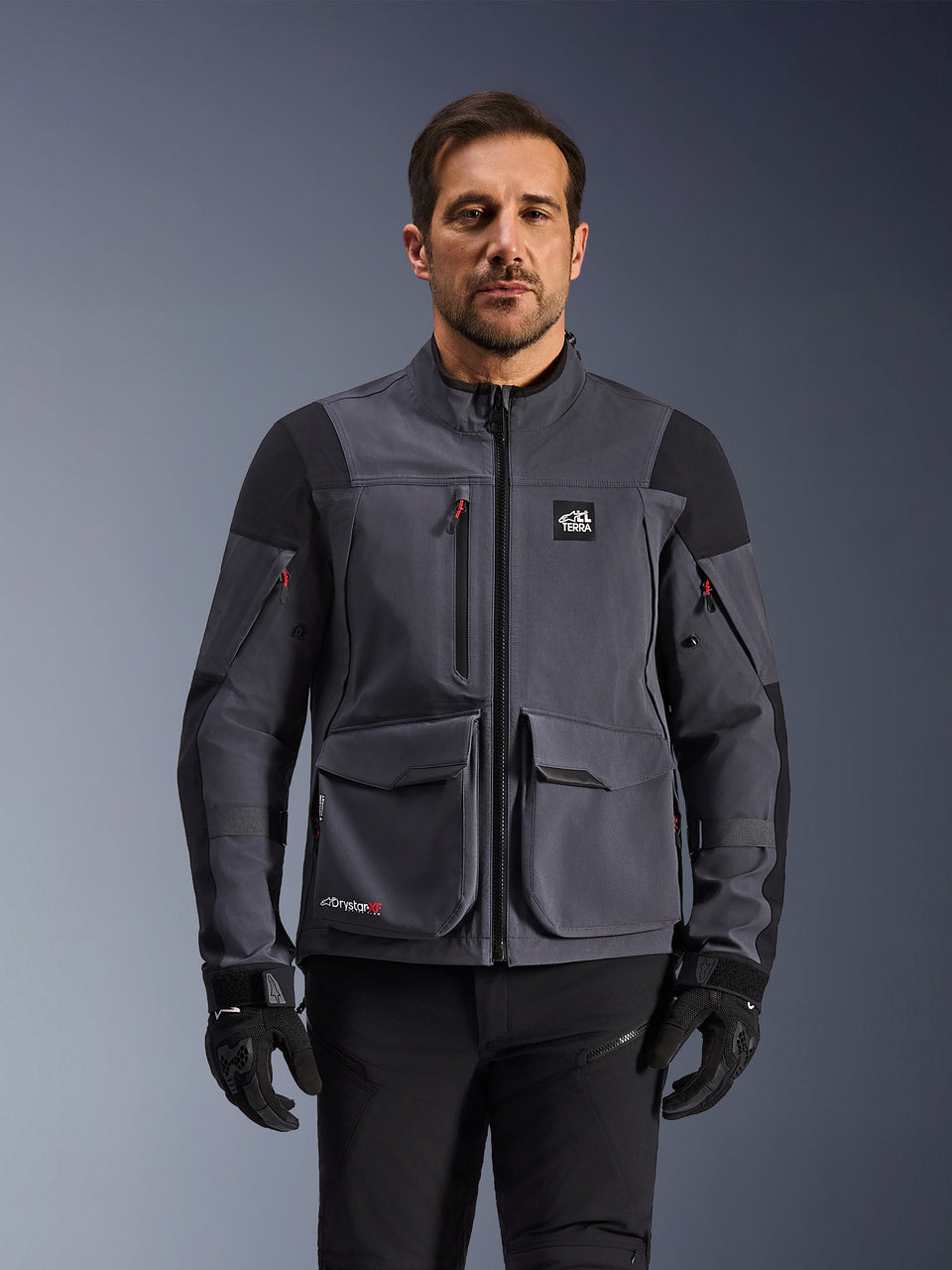 Alpinestars Maxdura Drystar®XF Laminierte Gepanzerte Schale, Adventure-Motorradjacke in Deep Gray und Schwarz, Vorderansicht am Modell, mit großen Cargotaschen, 'All Terra'-Brustlogo und Belüftungsreißverschlüssen, kombiniert mit schwarzen Motorradhandschuhen und -hosen.