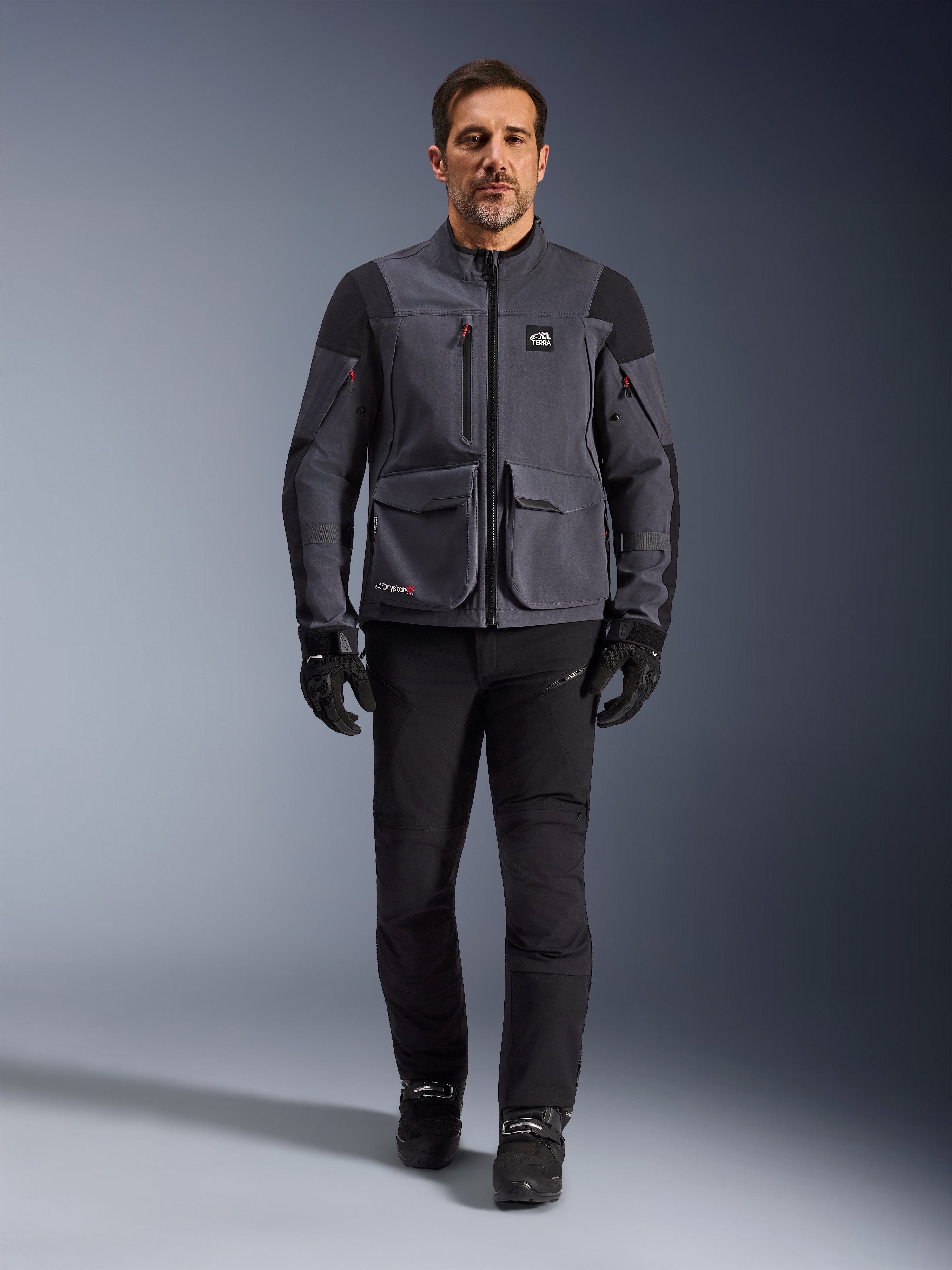 Maxdura Drystar®XF Laminierte Gepanzerte Jacke, Motorradjacke, Dunkelgrau-Schwarz, getragen von einem Modell mit schwarzer Hose und Handschuhen, mit großen Cargotaschen, roten Reißverschlussakzenten und All Terra-Branding, technische Adventure-Fahrausrüstung