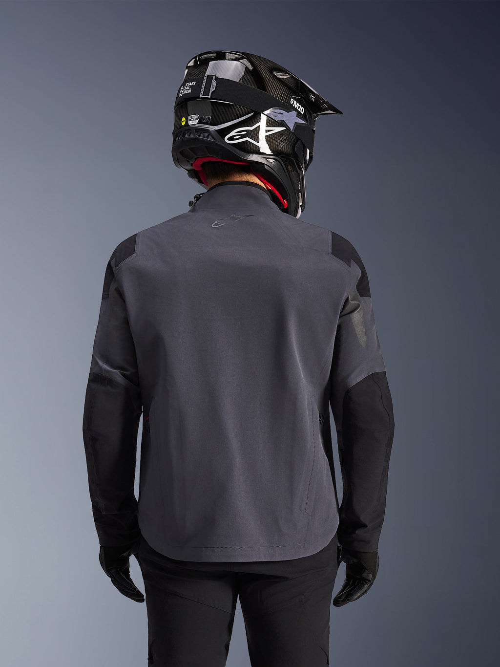 Maxdura Drystar®XF Laminierte Gepanzerte Jacke, Adventure-Motorradjacke, Dunkelgrau Schwarz, Rückansicht mit technischer Textilkonstruktion und verstärkten Panelen, getragen mit schwarzem Carbon-Helm, schwarzer Fahrhose und Handschuhen